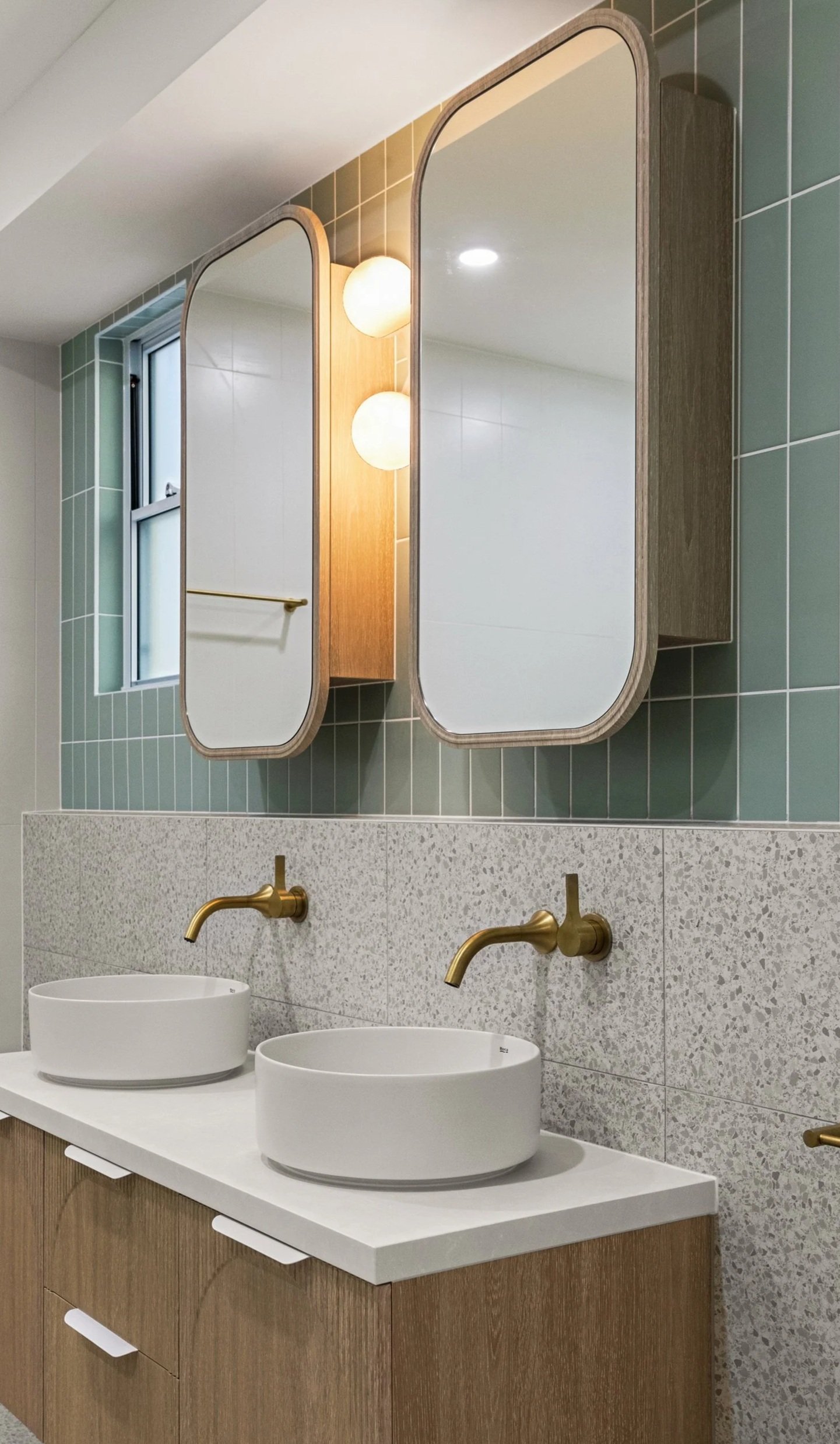Bathroom with two round vessel sinks on a white countertop, brass faucets, a tiled wall with rectangular mirrors, a window, a wooden cabinet, and modern lighting fixtures.