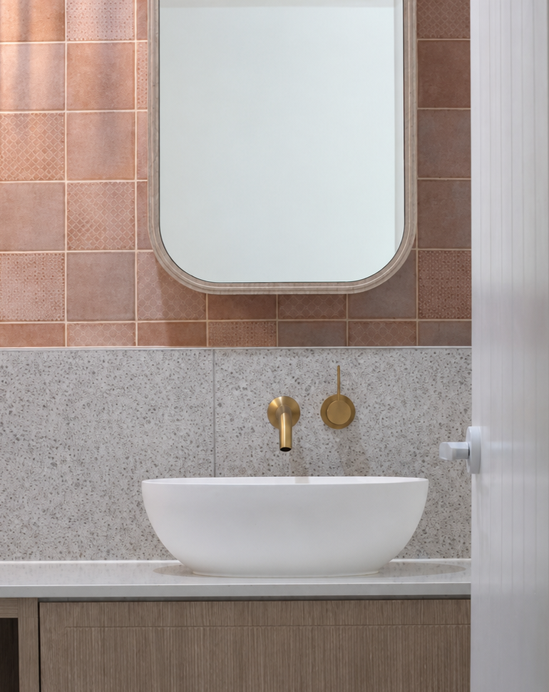 Bathroom sink with a white vessel basin, gold faucet, and a round gold wall fixture above it. A mirror with rounded corners is above the sink, with pink tiles on the wall behind and a textured grey tile below.