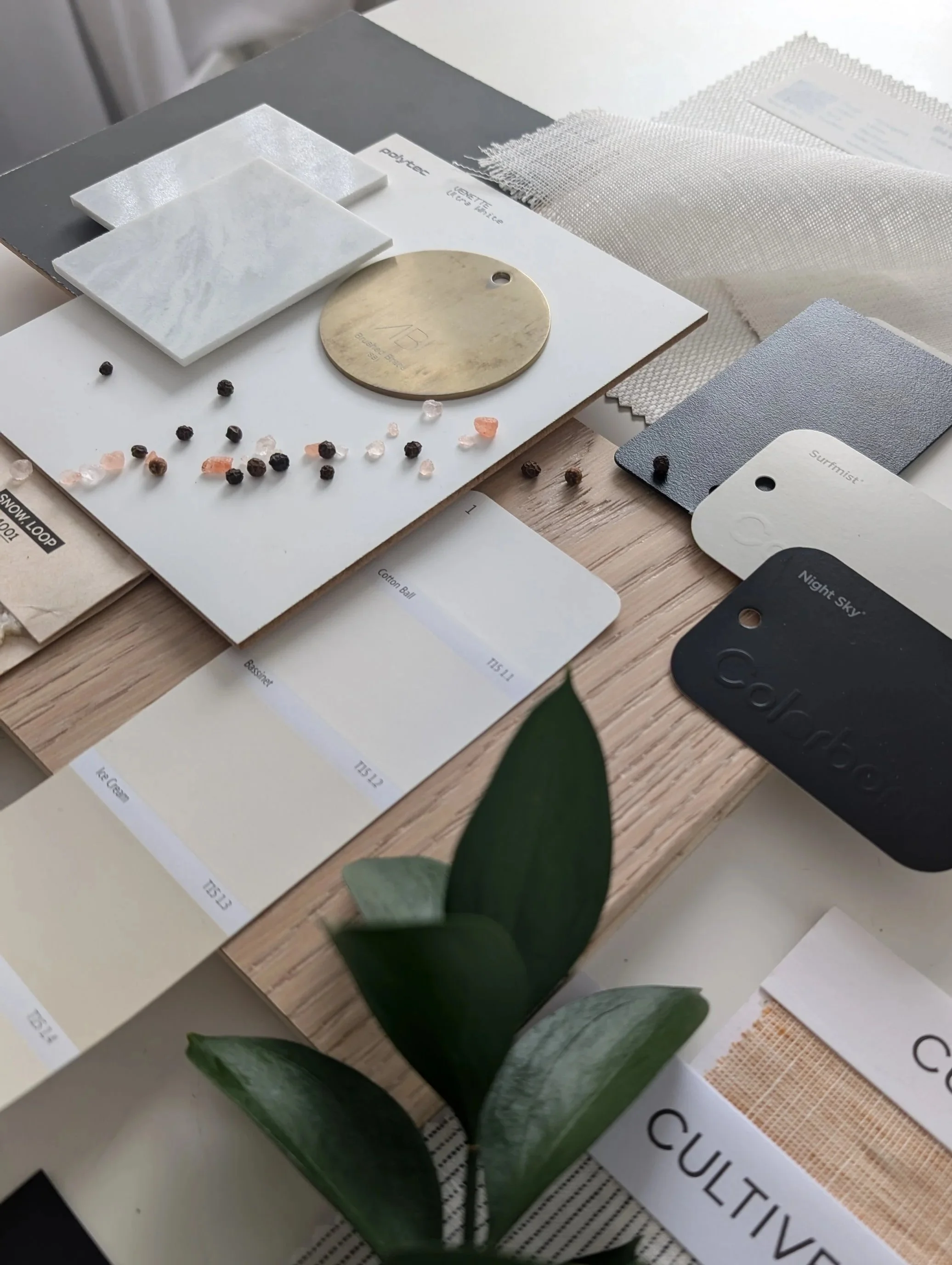 Close-up of interior design material samples, including tiles, fabrics, and color swatches, on a wooden surface with a plant in the foreground.