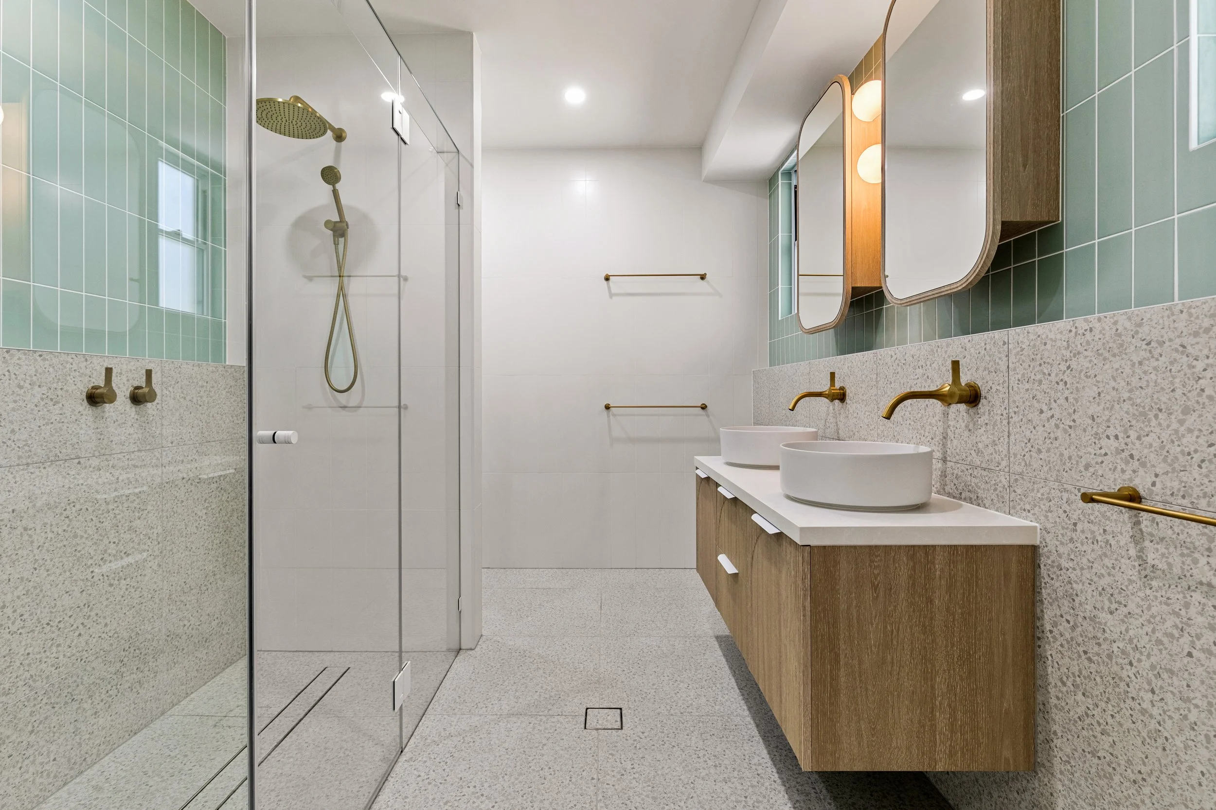 Modern bathroom featuring a walk-in shower with glass doors, a dual vanity with two white vessel sinks, gold fixtures, large mirrors with wooden framing, green tiled wall accents, and terrazzo-style flooring.