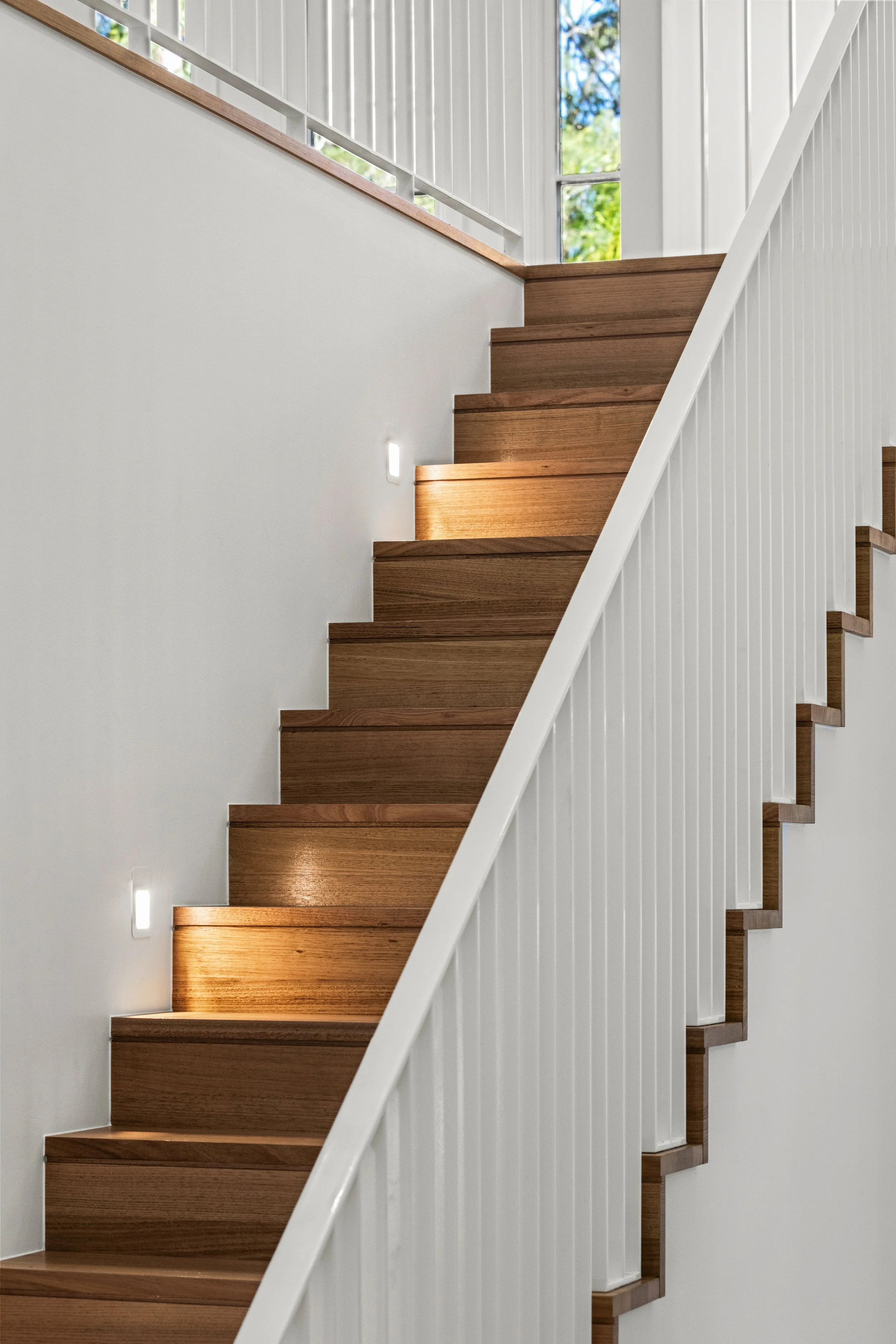 Modern staircase with wooden steps, white railing, and wall-mounted lights, leading up toward a large window showing trees outside.
