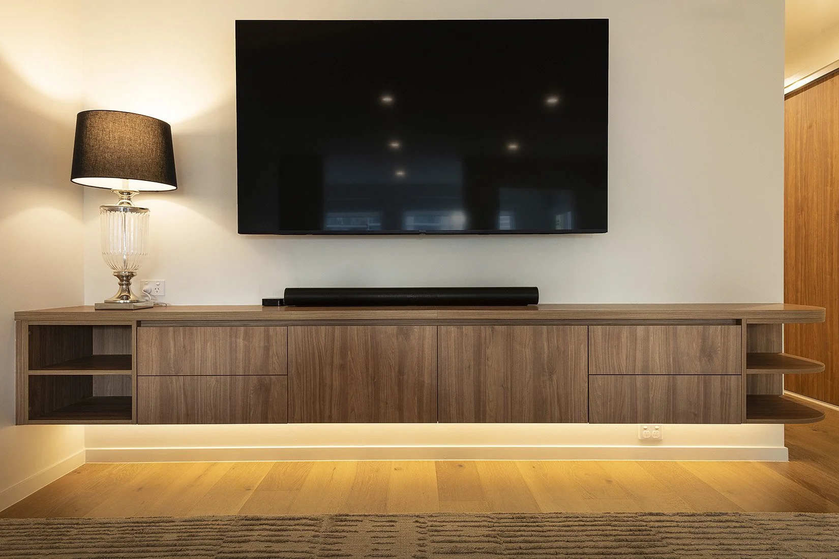 Living room entertainment center with a flat-screen TV, a soundbar below, a black table lamp with a glass base on the left, and a wooden cabinet with open shelves and drawers, illuminated by warm lighting.