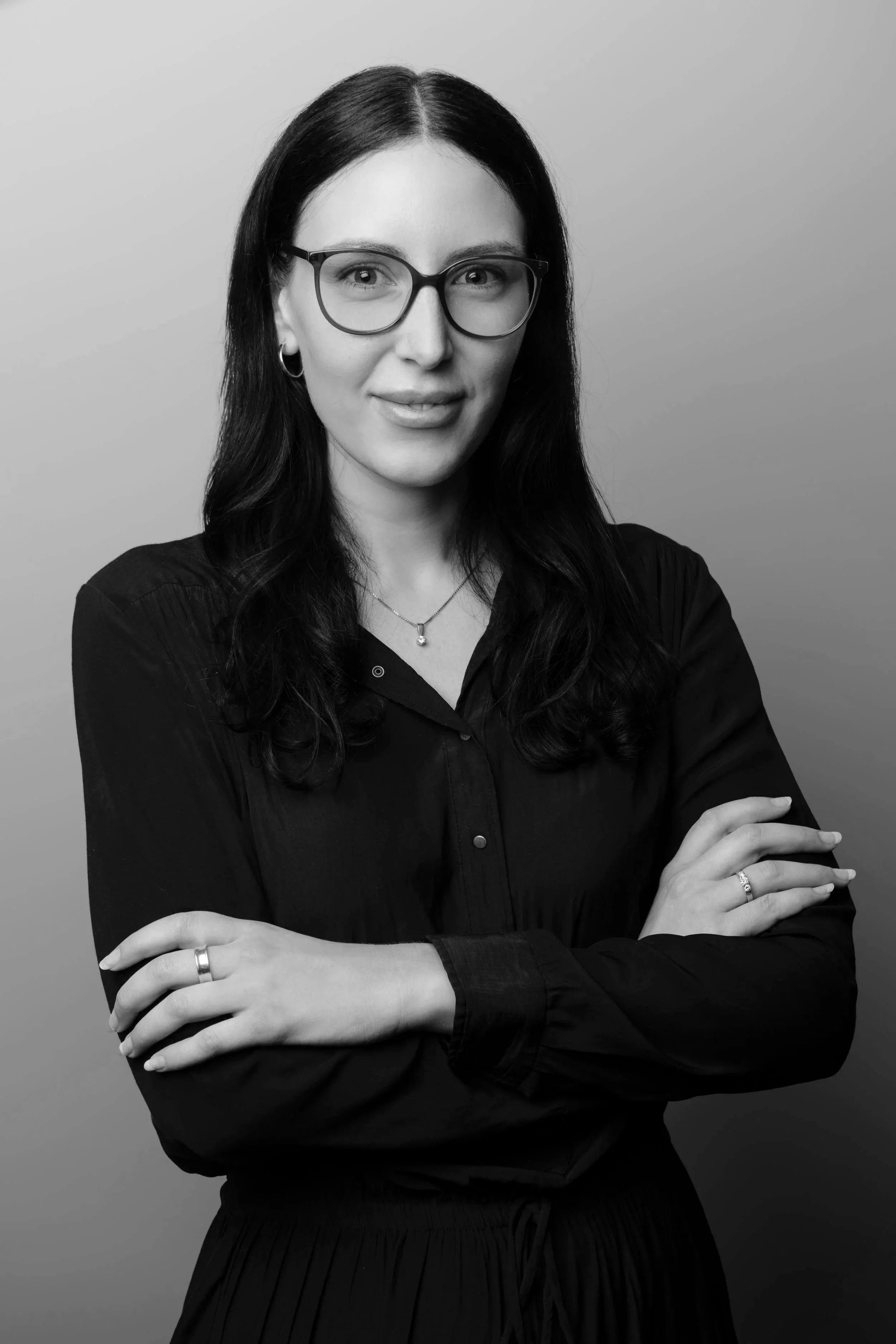 Black and white portrait of a woman with long dark hair, wearing glasses, earrings, and a necklace, crossed arms, smiling subtly against a plain background.