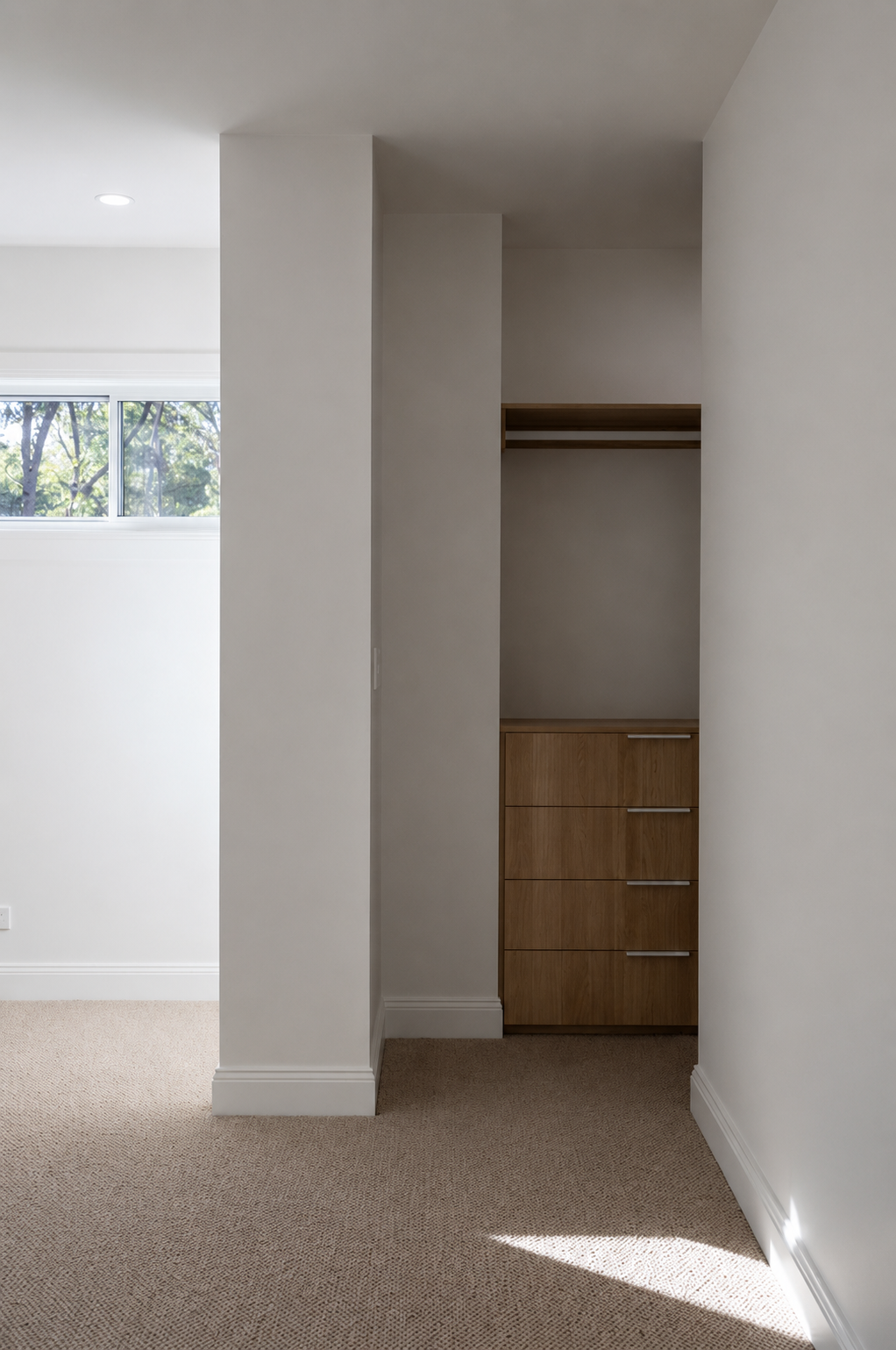 Interior view of a minimalist room with a small built-in wooden cabinet and drawers, beige carpet, off-white walls, and a window with trees outside.