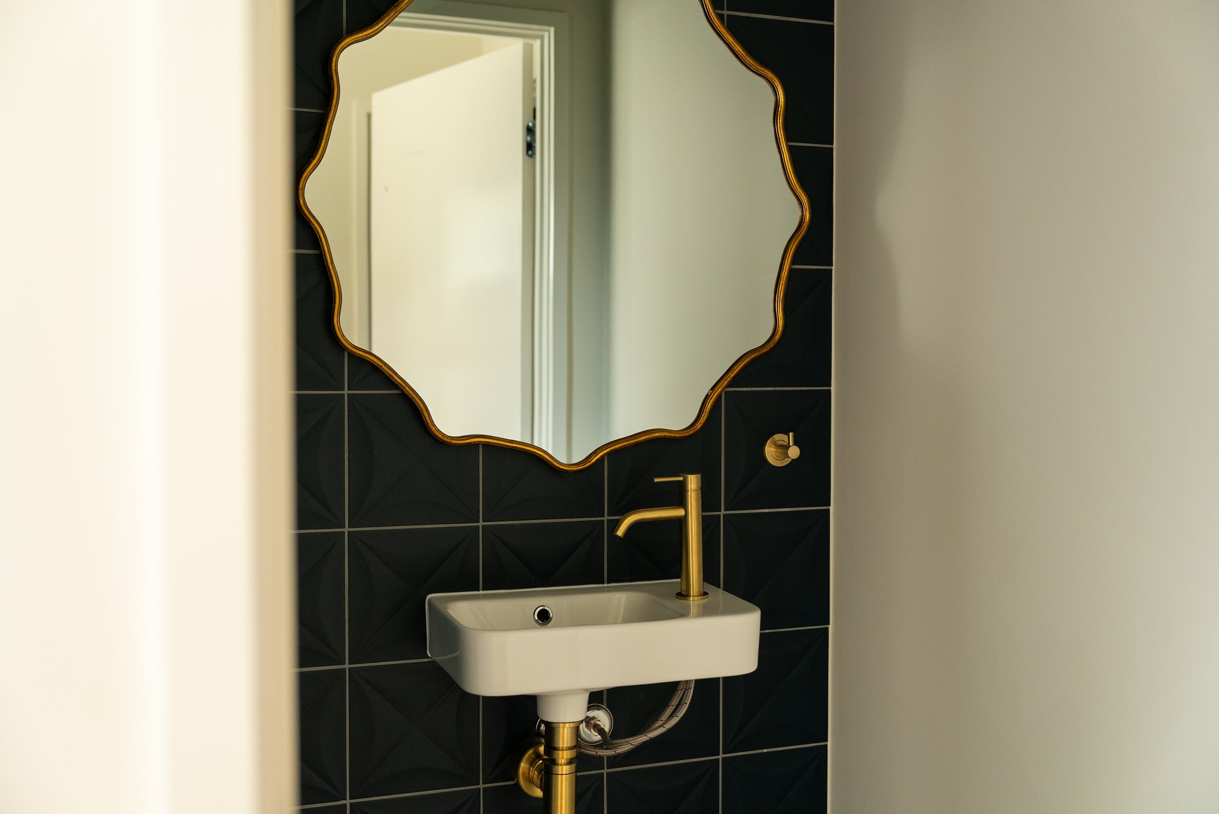 A bathroom with a decorative mirror, a white sink, and a gold faucet. The background features black tiles with geometric patterns and a white wall. There is a door reflected in the mirror.