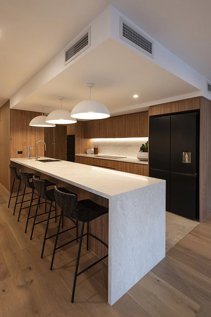 Modern kitchen with a white marble island, black bar stools, wooden cabinets, and a black refrigerator. Three white pendant lights hang above the island.