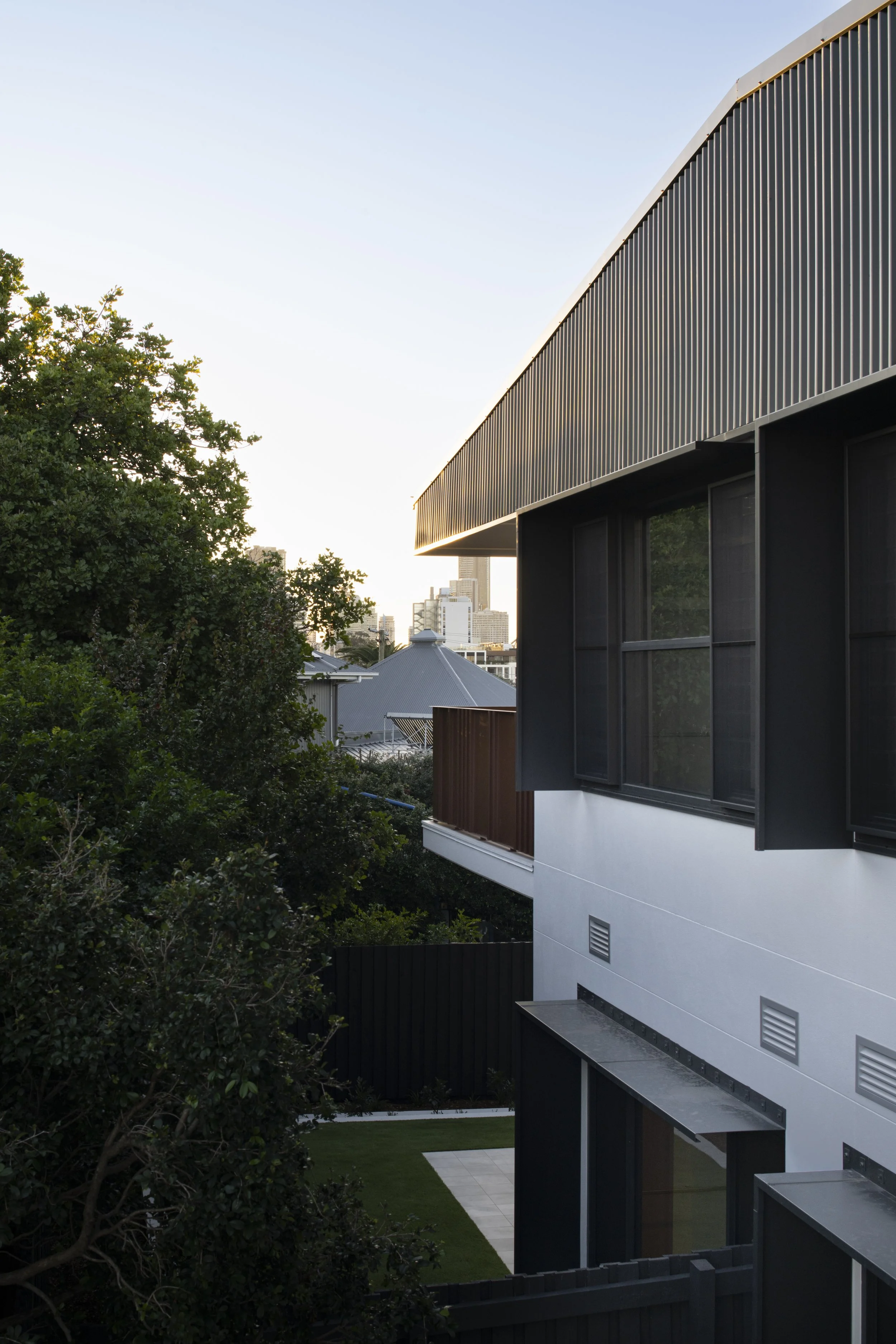 Modern residential building with large windows and a metal cladding exterior, surrounded by greenery and with a city skyline in the background during sunset.
