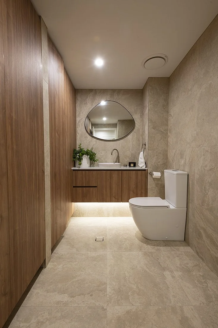 Modern bathroom with beige tiled floors and walls, wooden cabinets, a round mirror above the sink, a white toilet, and lighting beneath the sink with a small green plant on the cabinet.
