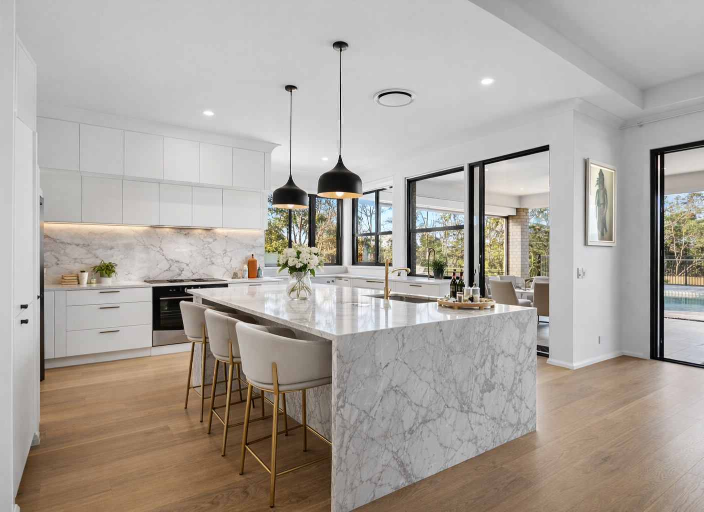 Modern kitchen with white cabinetry, marble countertops, and a large island with seating, black pendant lights, and windows overlooking a backyard with trees and a pool.