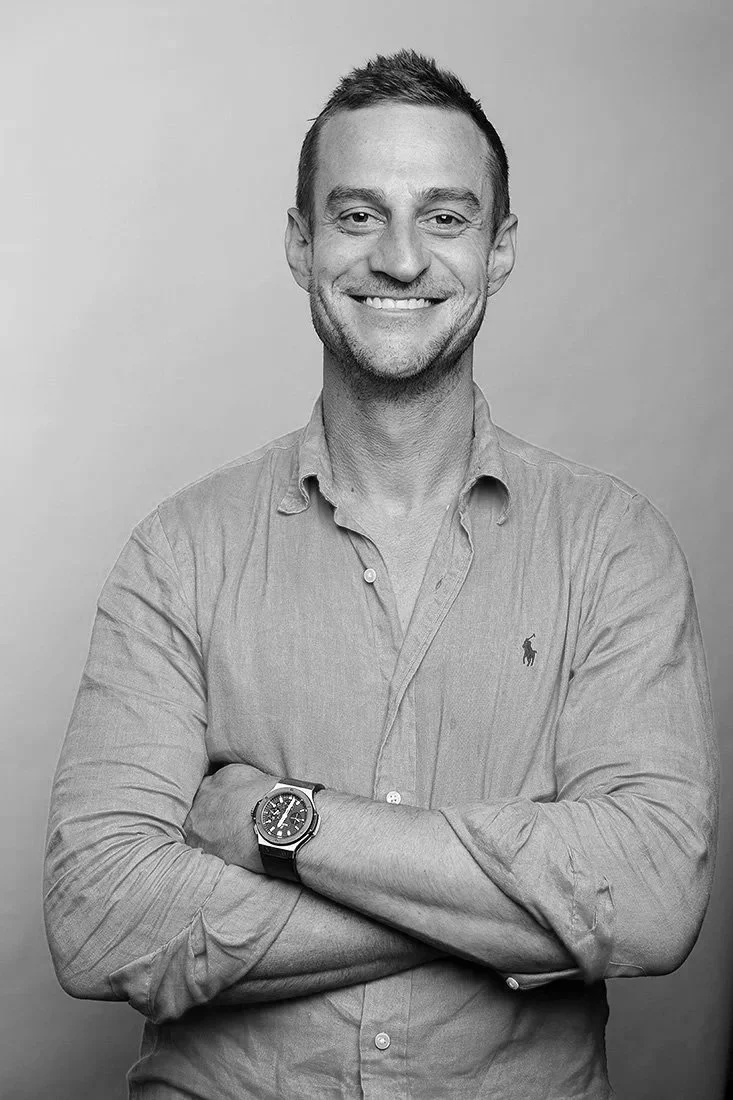 Black and white portrait of a smiling man with short hair, wearing a button-up shirt with the sleeves rolled up, crossed arms, and a watch on his left wrist.