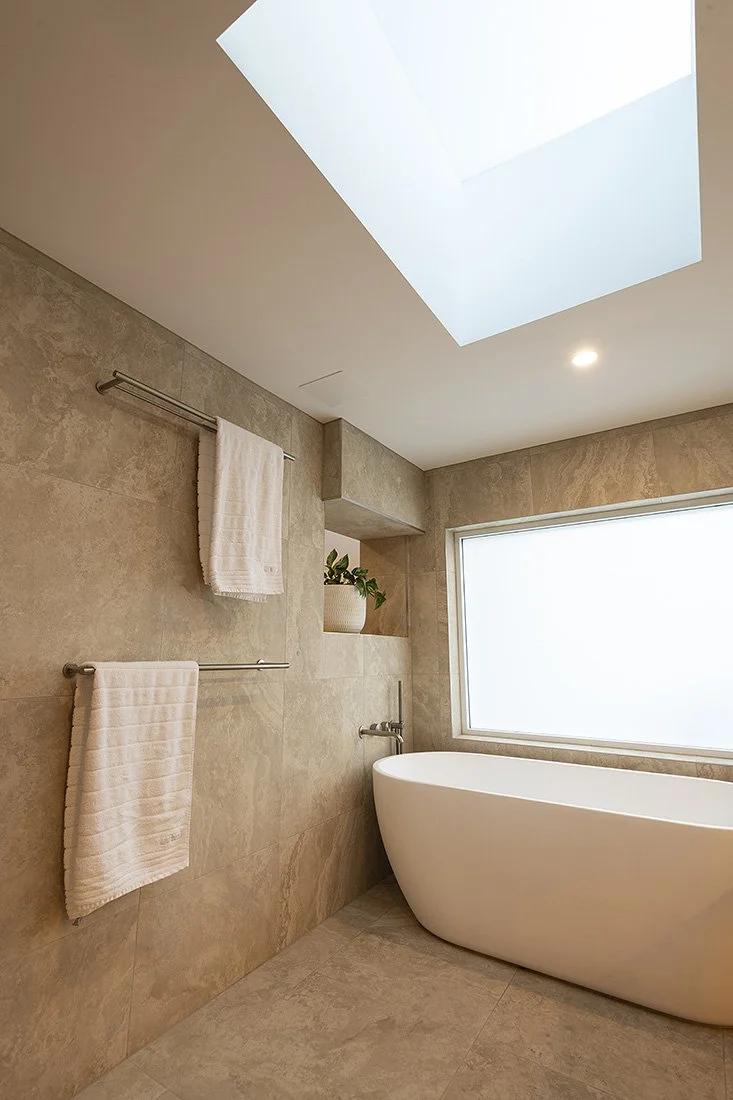 Modern bathroom with beige tiled walls and floor, a large frosted window, a white freestanding bathtub, built-in shelves with a potted plant, and towel racks with white towels. Recessed ceiling lights and a skylight provide natural and artificial lig