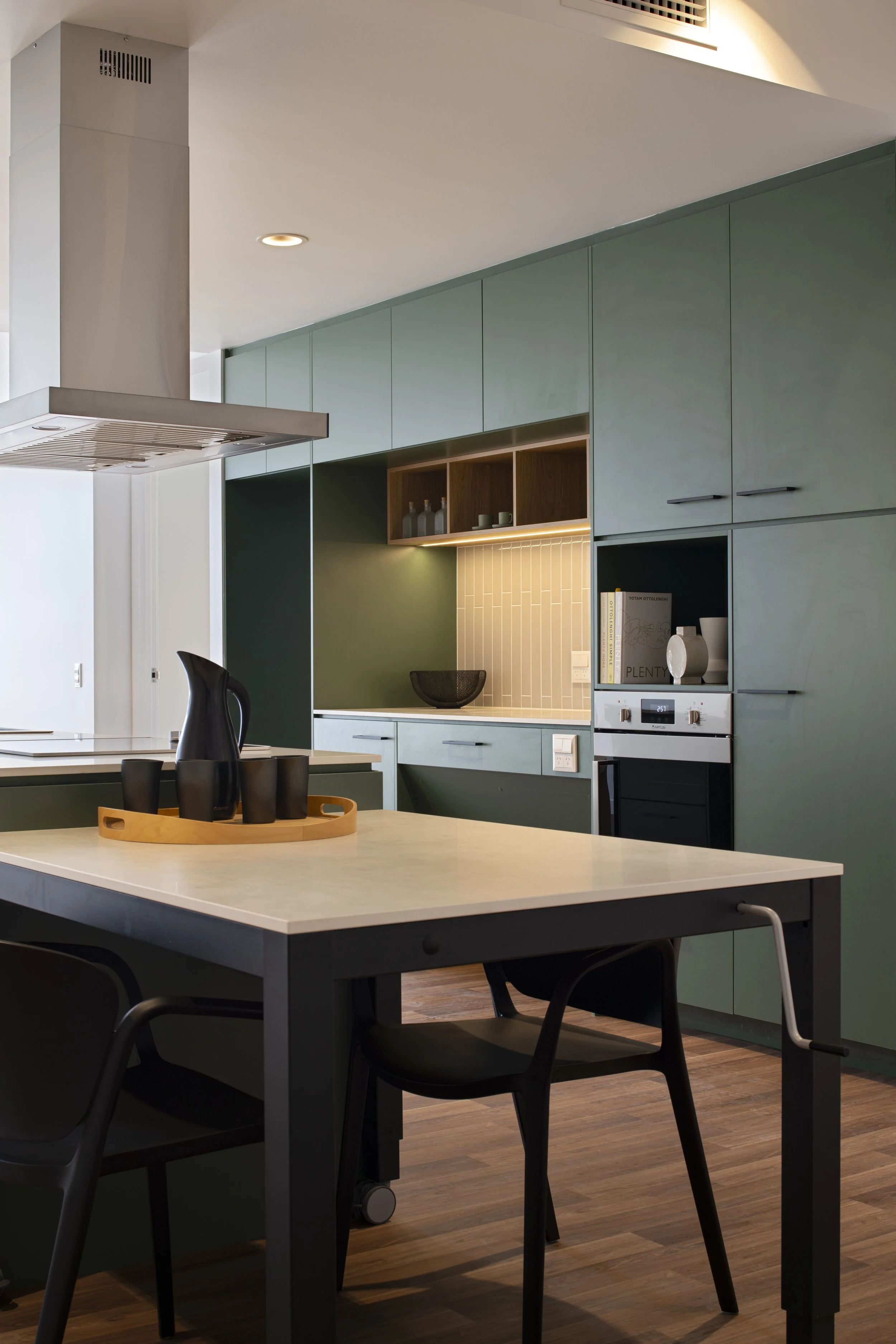 Modern kitchen with gray cabinets, a cream countertop, and a white and wood backsplash. There is a black pitcher and cups on a wooden tray on the island, and black chairs around the table.