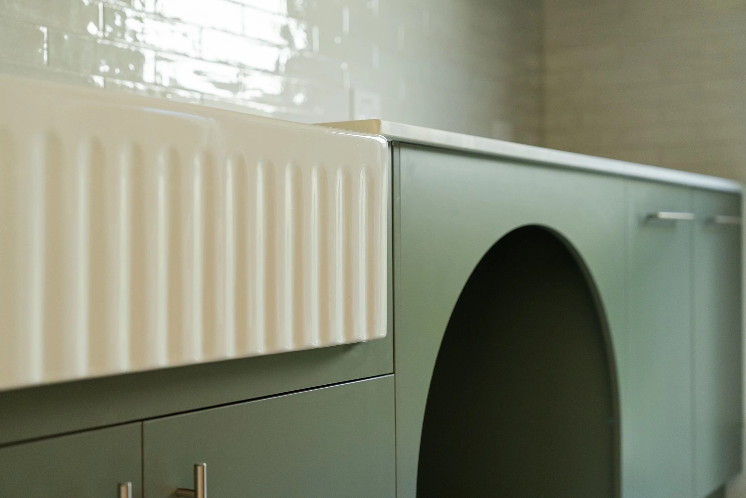 Close-up of a modern washing machine in a muted gray-green color in a laundry room with a beige radiator on the left and a tiled wall in the background.