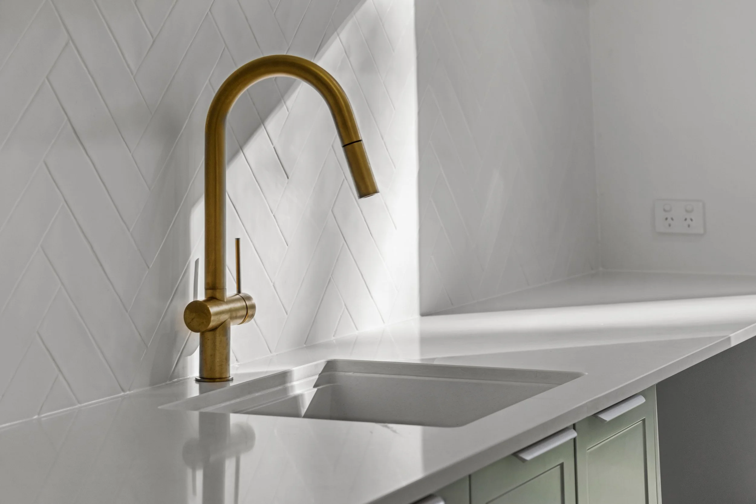 Close-up of a modern kitchen sink with a gold-colored faucet, white tiled backsplash, and a white countertop with cabinetry below.