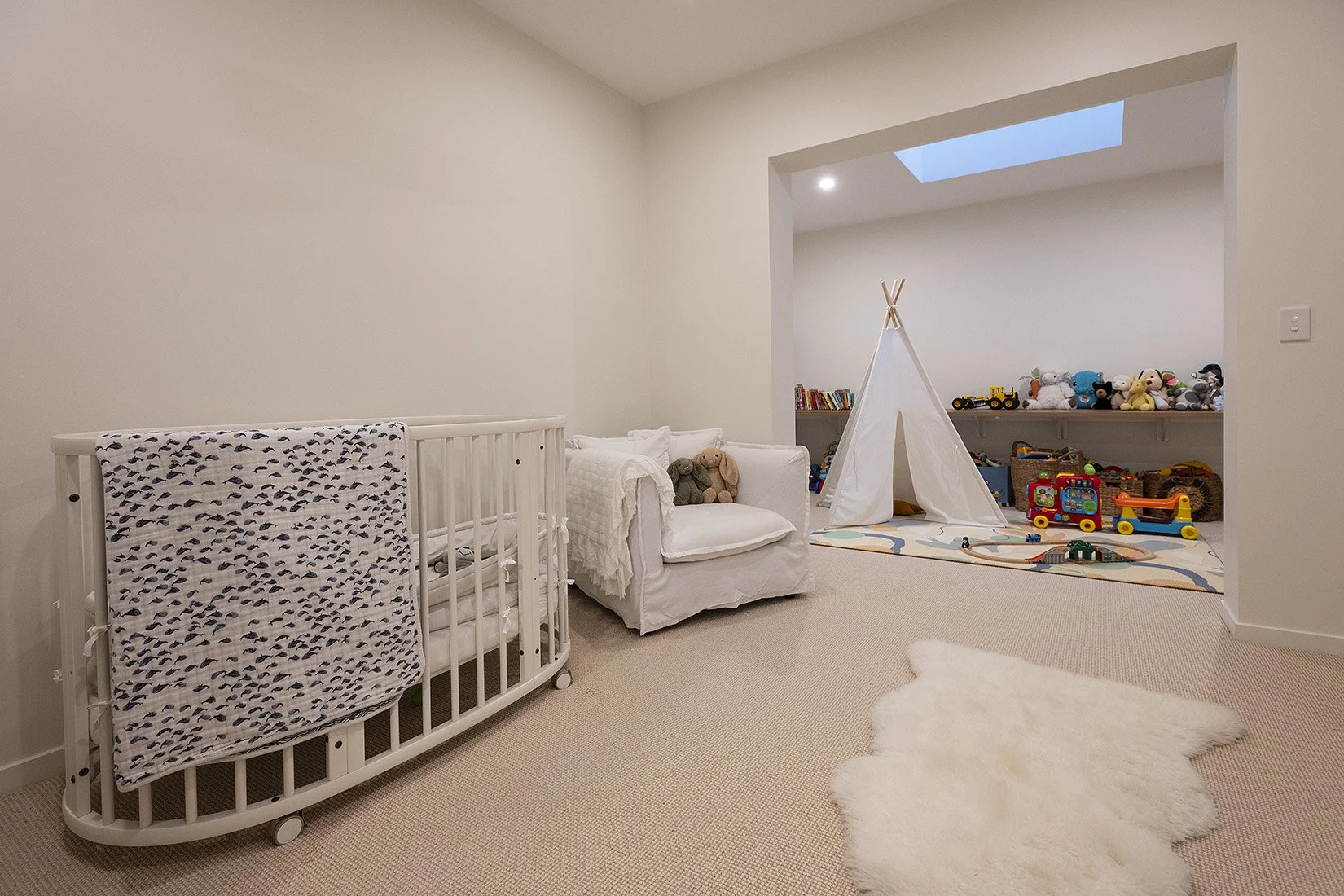 A children's playroom with white walls and beige carpet. Features include a white crib, a white sofa with stuffed animals, a children's teepee tent, toys on shelves, a colorful play rug, and plush toys displayed on a shelf.