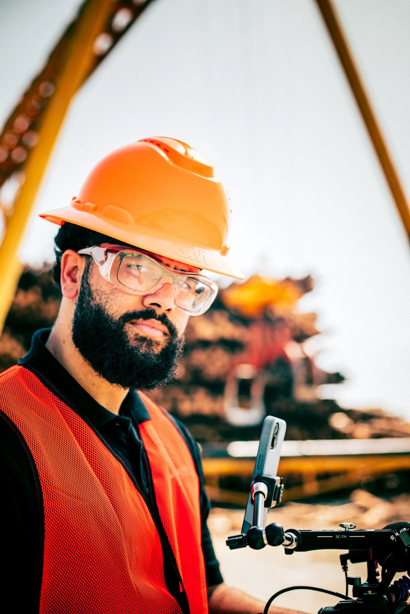 A male construction worker with a beard and glasses, wearing an orange safety vest and a yellow hard hat, operating a drone or remote-controlled device outdoors.