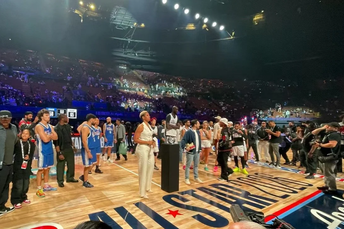 A basketball court with players, coaches, and officials gathered near the center. The stands are filled with spectators, and the arena is brightly lit, creating a lively atmosphere.