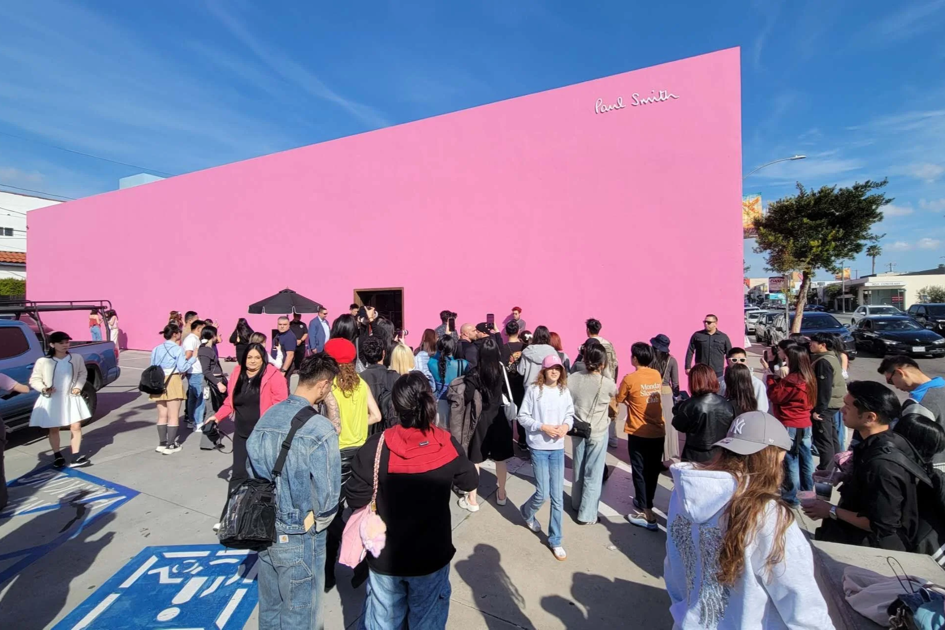 A crowd of diverse people gathered outside a pink building with Paul Smith written on it. The scene is sunny with a blue sky, and there are cars parked nearby.