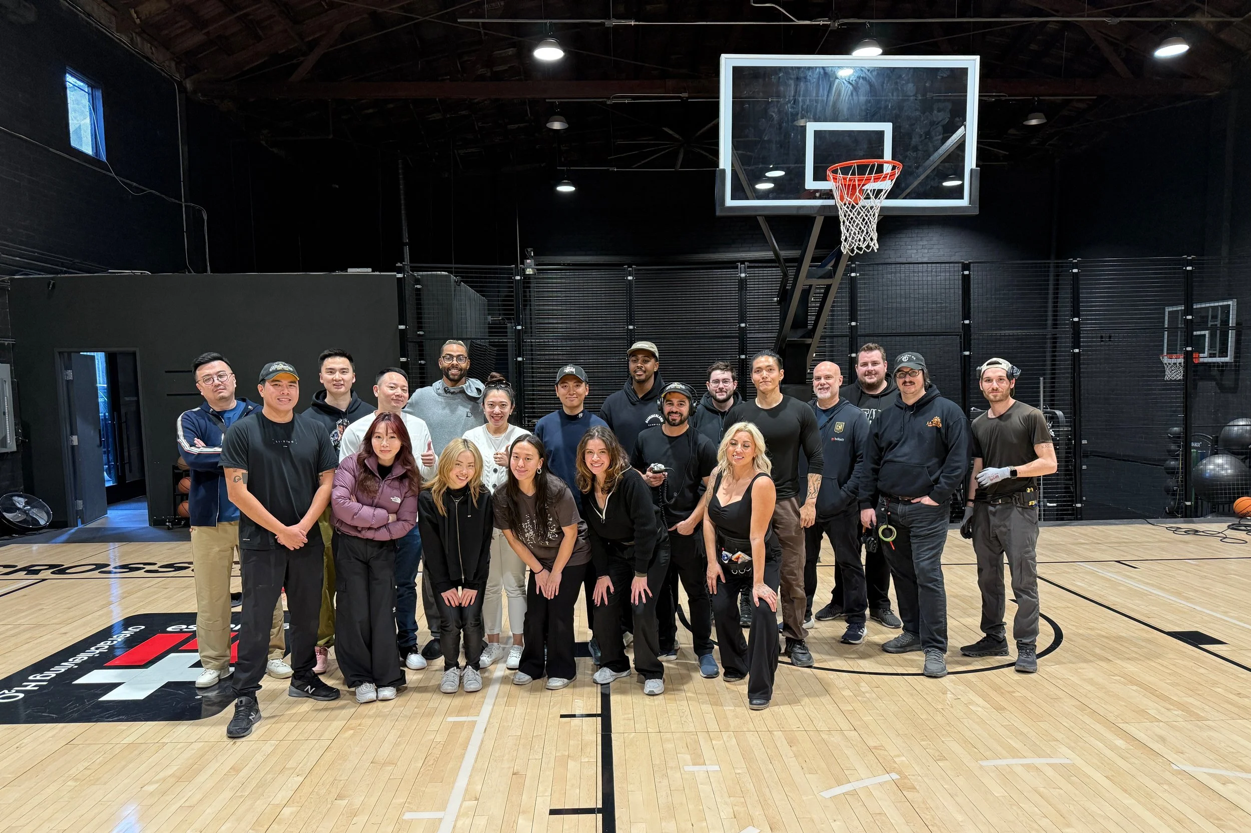 A group of diverse people posing on a basketball court inside a gym with black walls and high ceiling, smiling and standing in front of a basketball hoop.