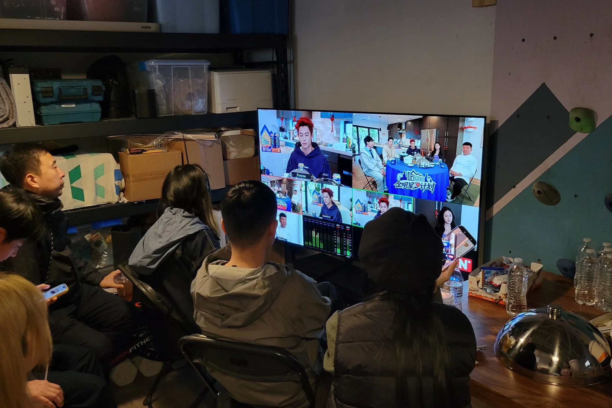 People watching a television screen displaying multiple camera angles of a panel discussion or interview in a room with cluttered shelves and various items on a table, including water bottles and snacks.