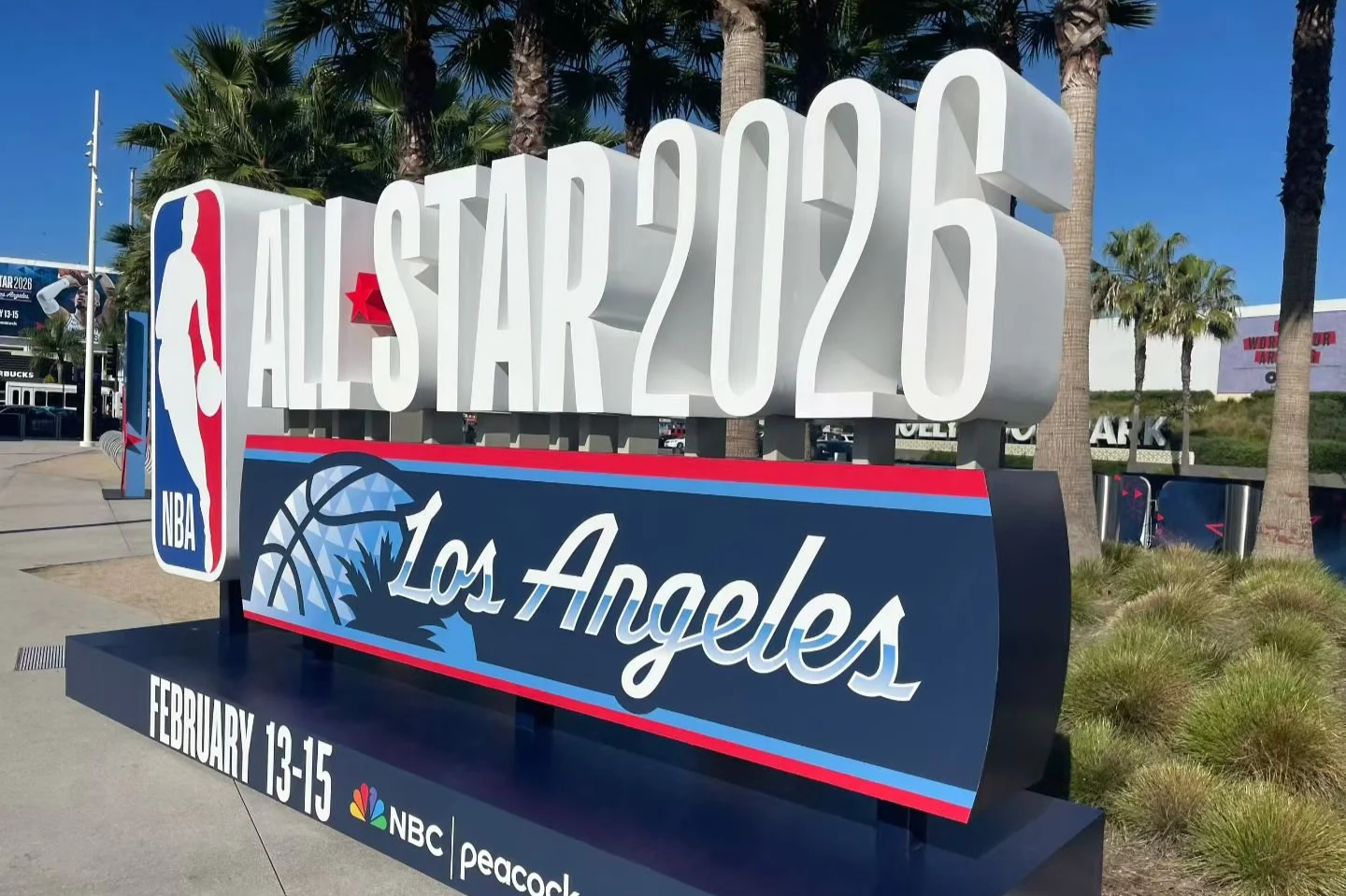 Outdoor sign for the NBA All-Star 2026 event in Los Angeles, featuring the NBA logo and event details, with palm trees and a clear blue sky in the background.