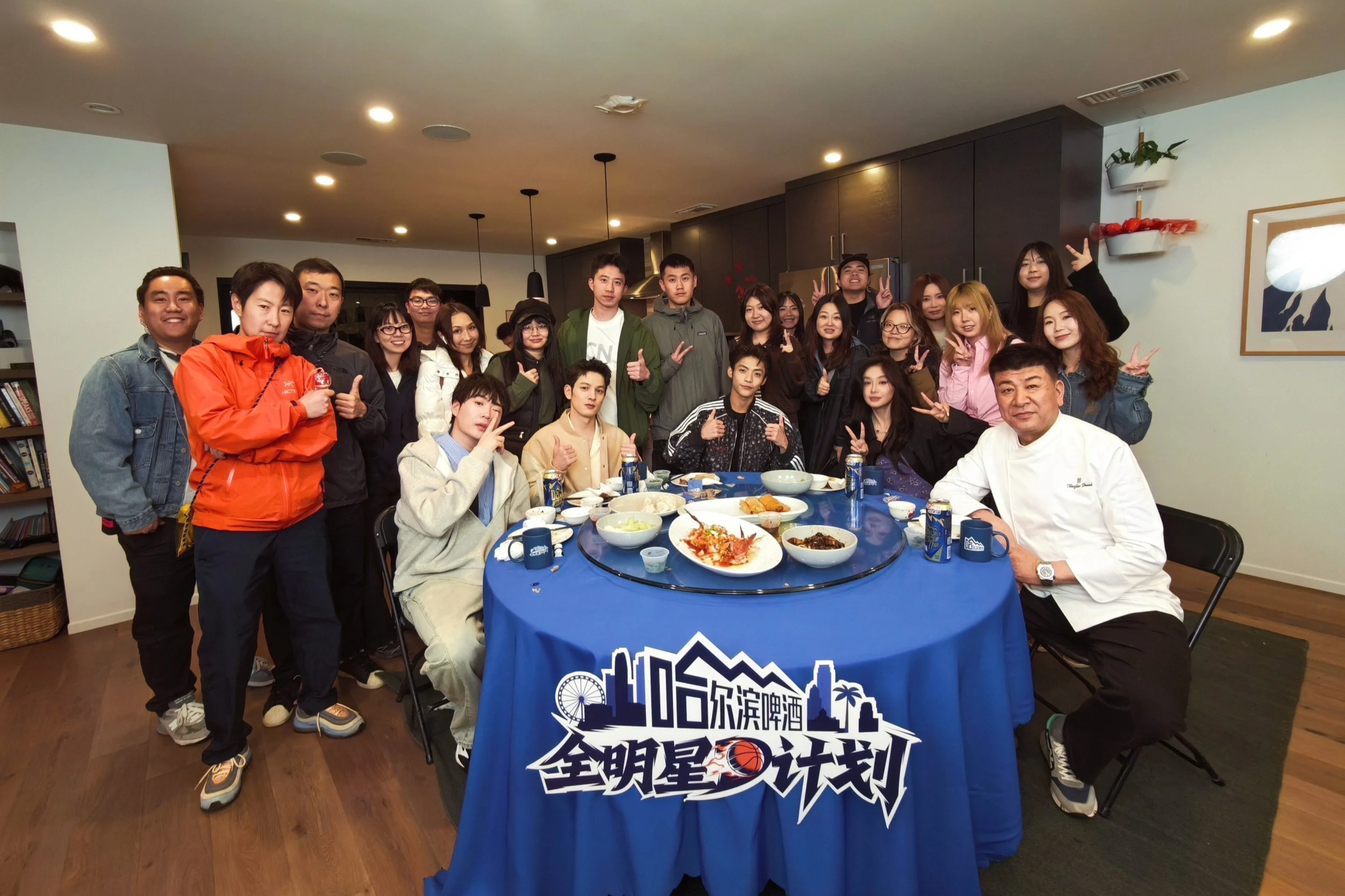 A large group of young adults and an older man, likely a chef, gathered around a table with various dishes, in a modern indoor setting, posing for a group photo with many making peace signs or thumbs up.