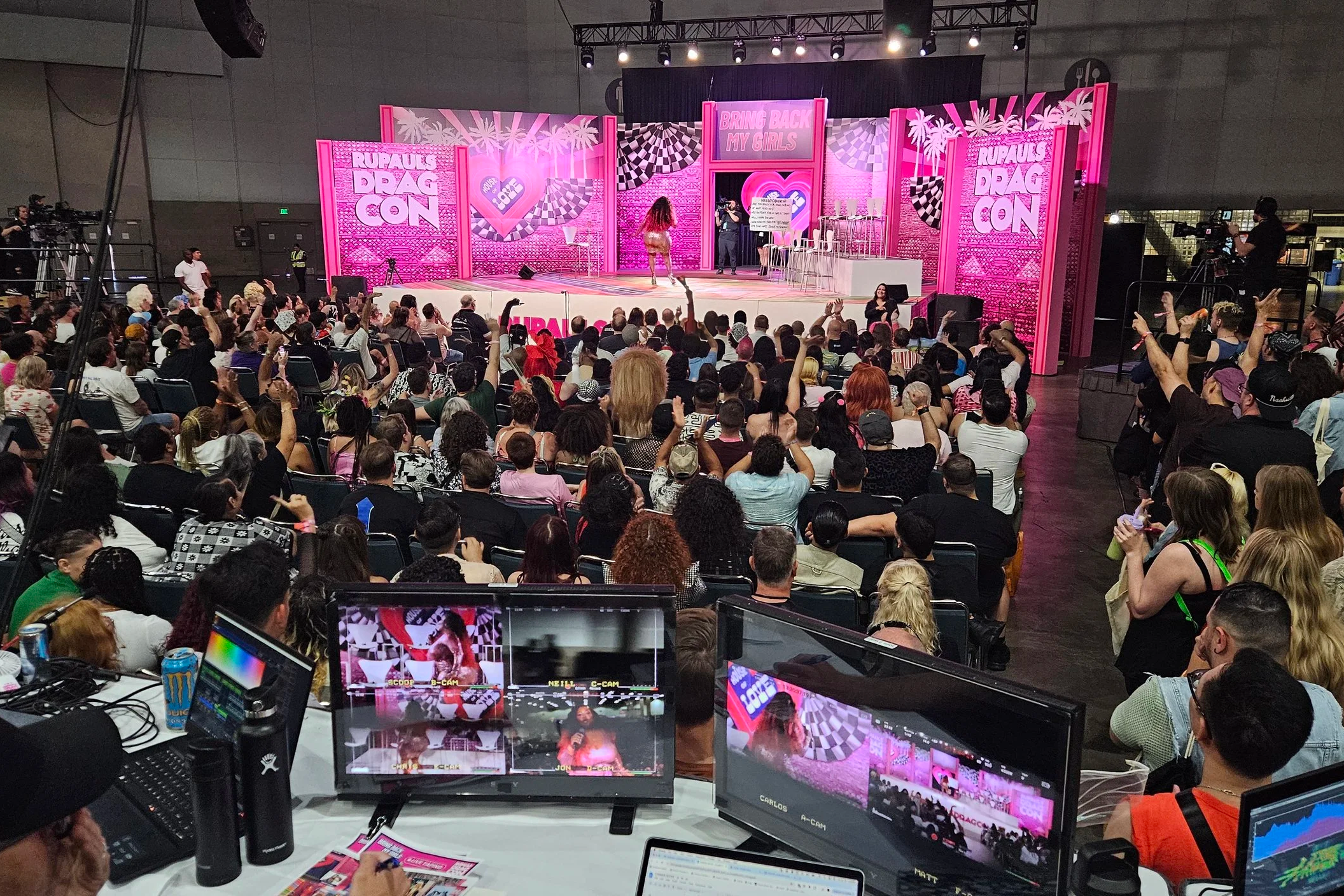 A stage at RuPaul's DragCon with pink themed decorations and the sign 'Bring Back My Girls' in the background. There are many attendees seated and cheering in front of the stage.