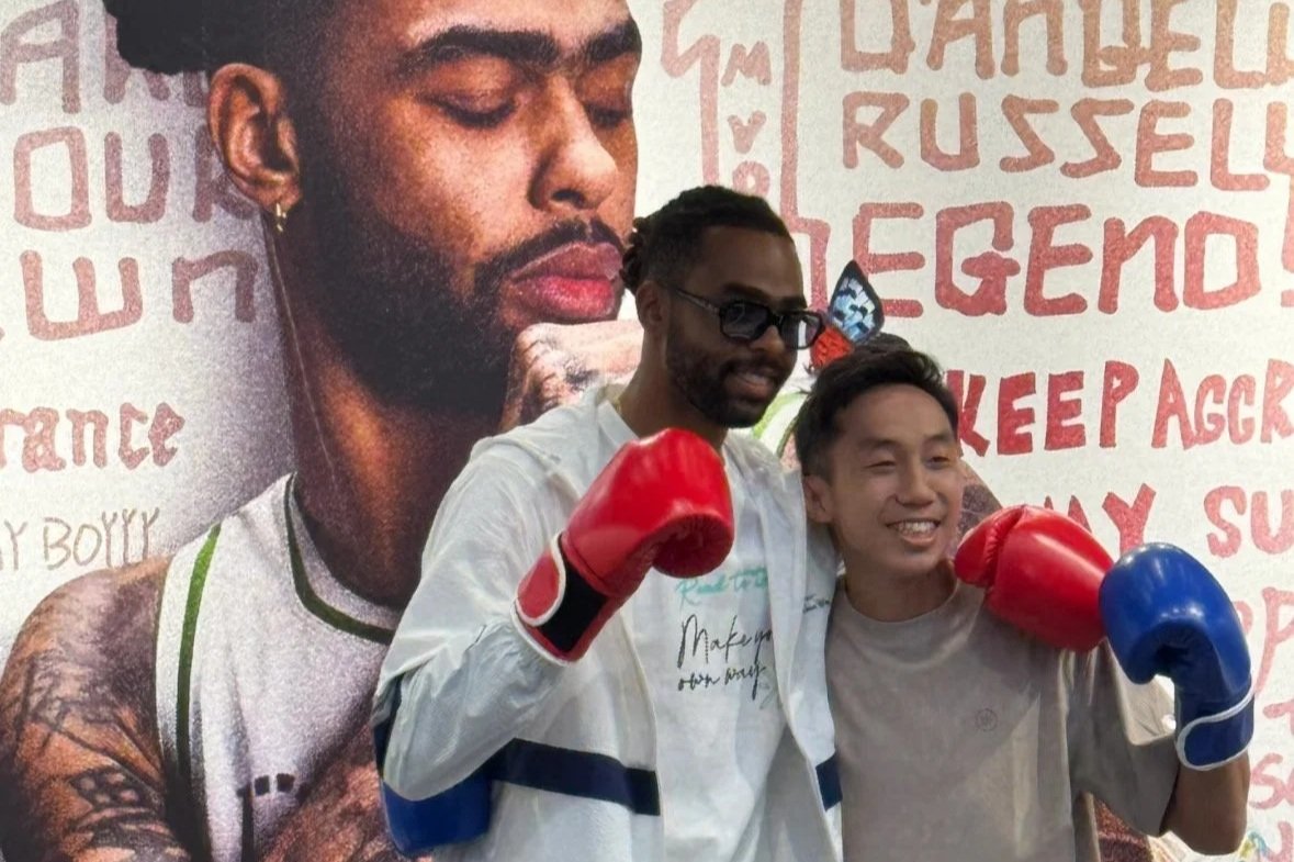 Two young men wearing boxing gloves, one with red gloves and the other with blue gloves, posing in front of a large portrait of a man with tattoos and a beard. The background has red text and drawings.