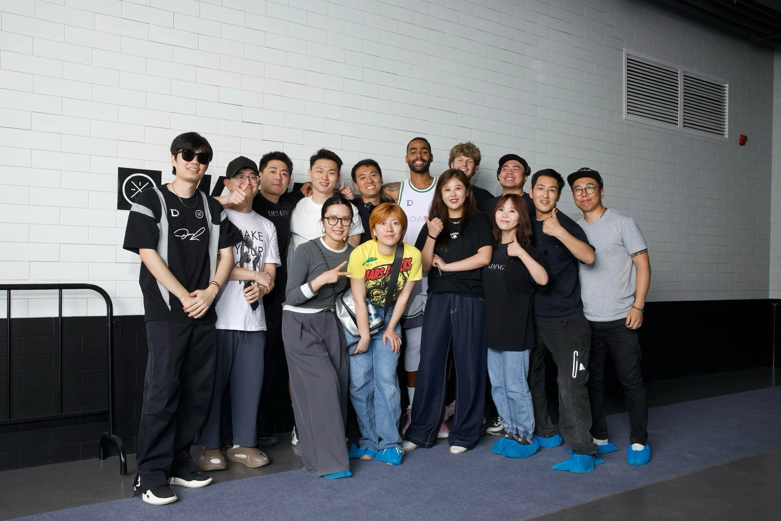 Group of diverse people standing together indoors against a white brick wall, some with arms around each other, wearing casual clothing and smiling.