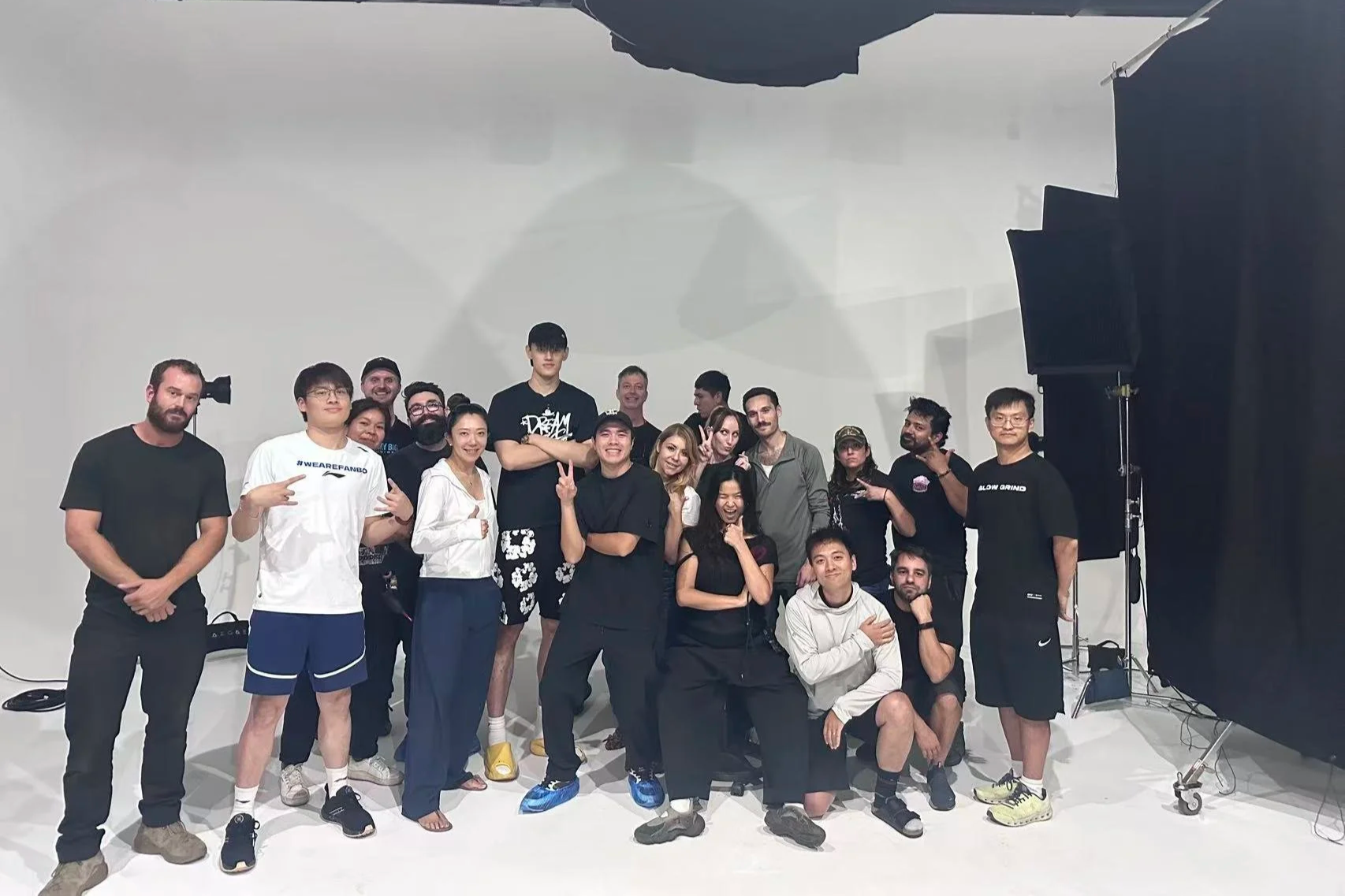 A group of diverse people posing together in a photography studio with a plain white background. Some are standing, some kneeling, and some sitting, smiling and making various gestures, with studio equipment visible on the sides.