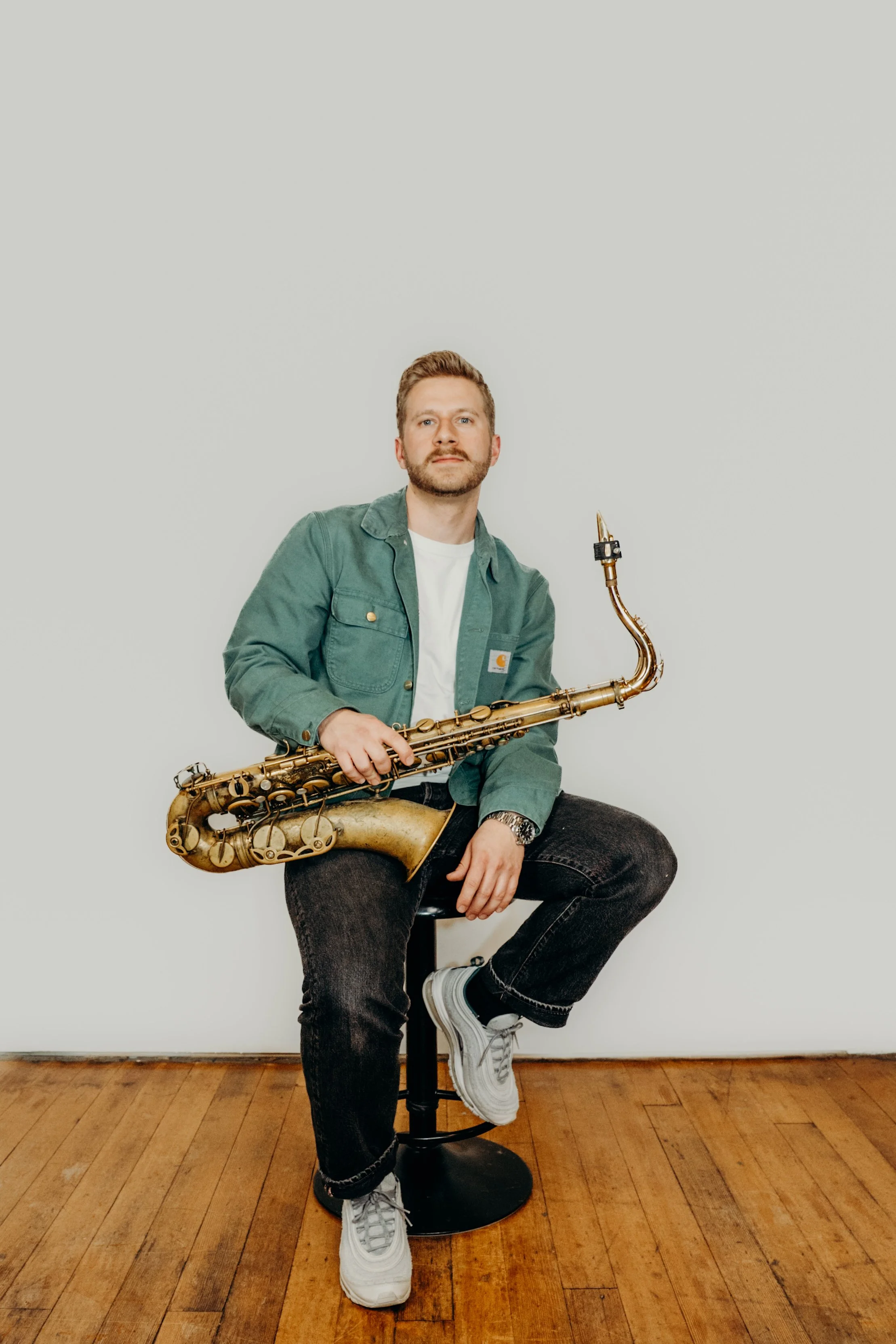 A man with a beard and light brown hair, dressed in a green jacket and white t-shirt, sitting on a stool holding a gold-painted saxophone, against a plain white background and wooden floor.