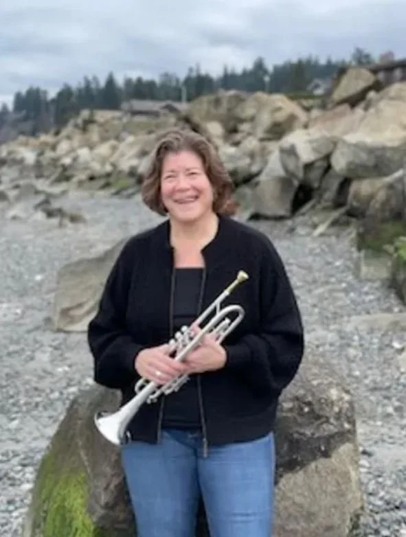 A woman smiling and holding a trumpet outdoors on a rocky beach with large rocks and trees in the background.