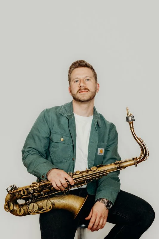 A man with a beard and short hair, wearing a green Carhartt jacket and white T-shirt, sitting and holding a saxophone against a plain white background.