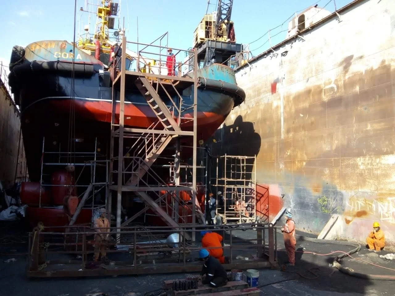 Trabajo en un dique de reparación de un buque industrial con varias personas en casco y utilizando equipo de protección, estructuras de andamios en el casco y herramientas en el suelo.