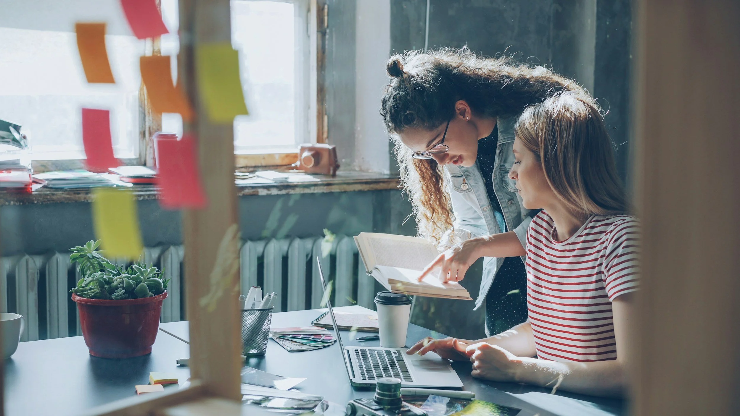 Two women collaborating in a bright, cozy office with natural light, surrounded by plants, sticky notes, a laptop, and various office supplies.