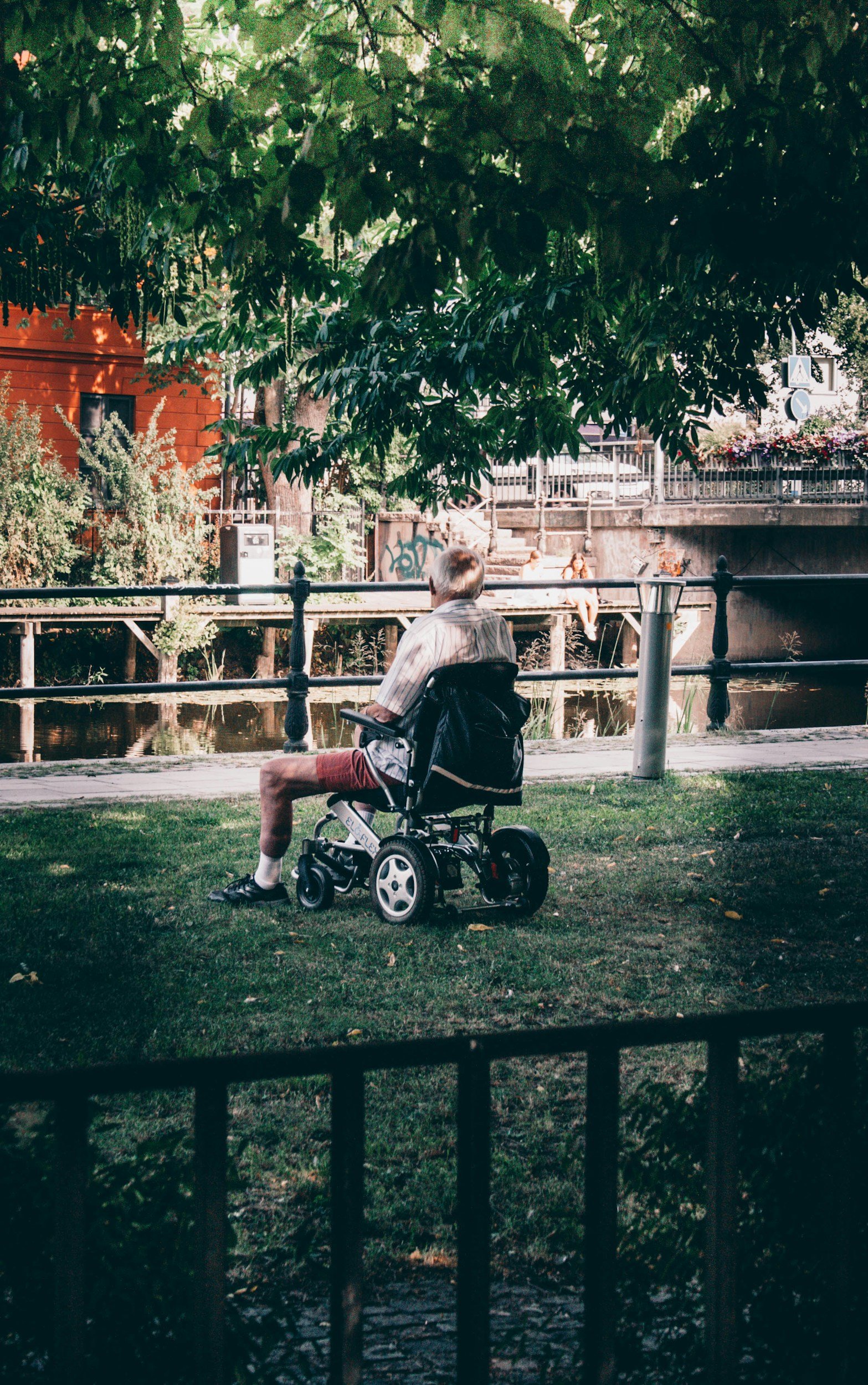 An elderly man sitting on a mobility scooter on a grassy area near a river in a park, with trees, a wooden fence, and a bridge with pedestrians in the background.