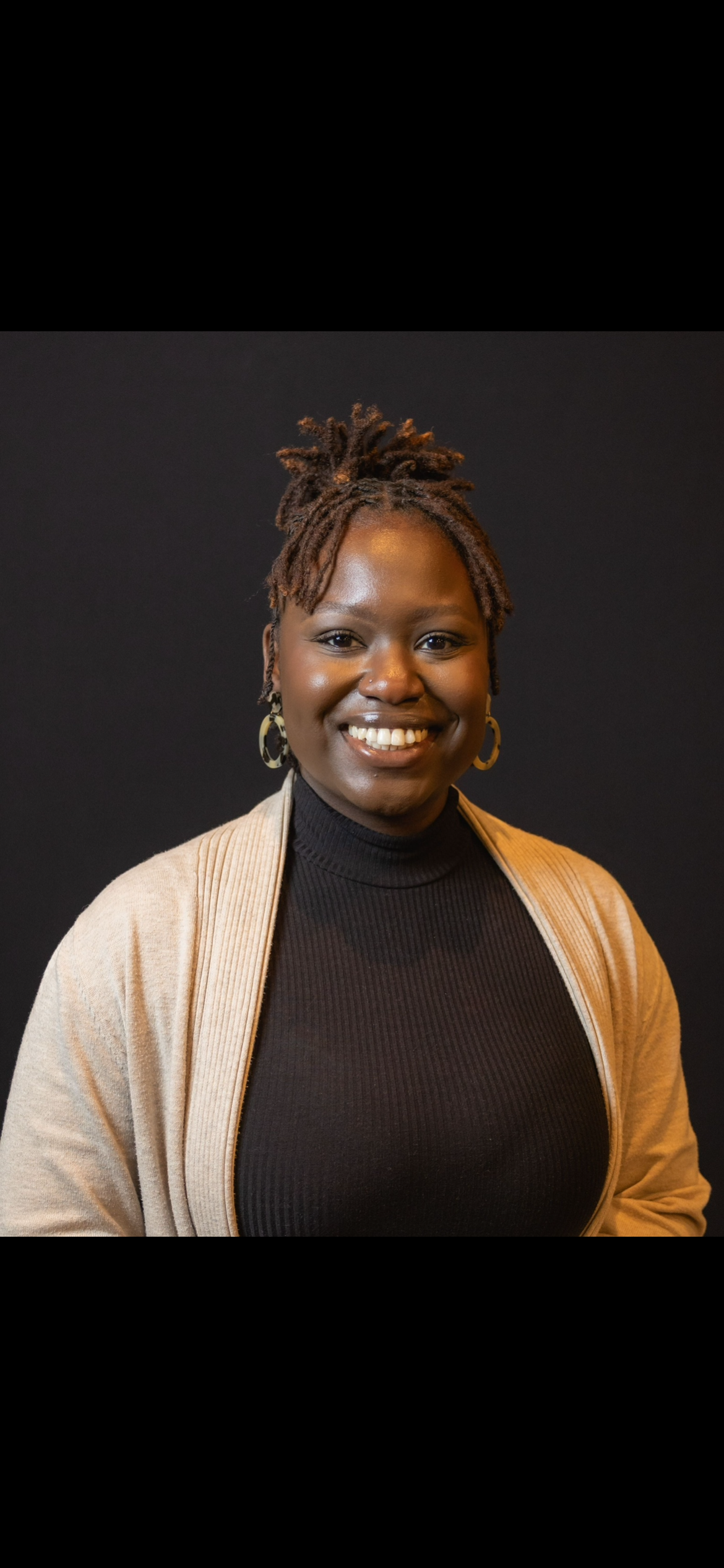Portrait of a smiling Black woman with dreadlocks, wearing a black turtleneck, gold earrings, and a beige cardigan, against a black background.