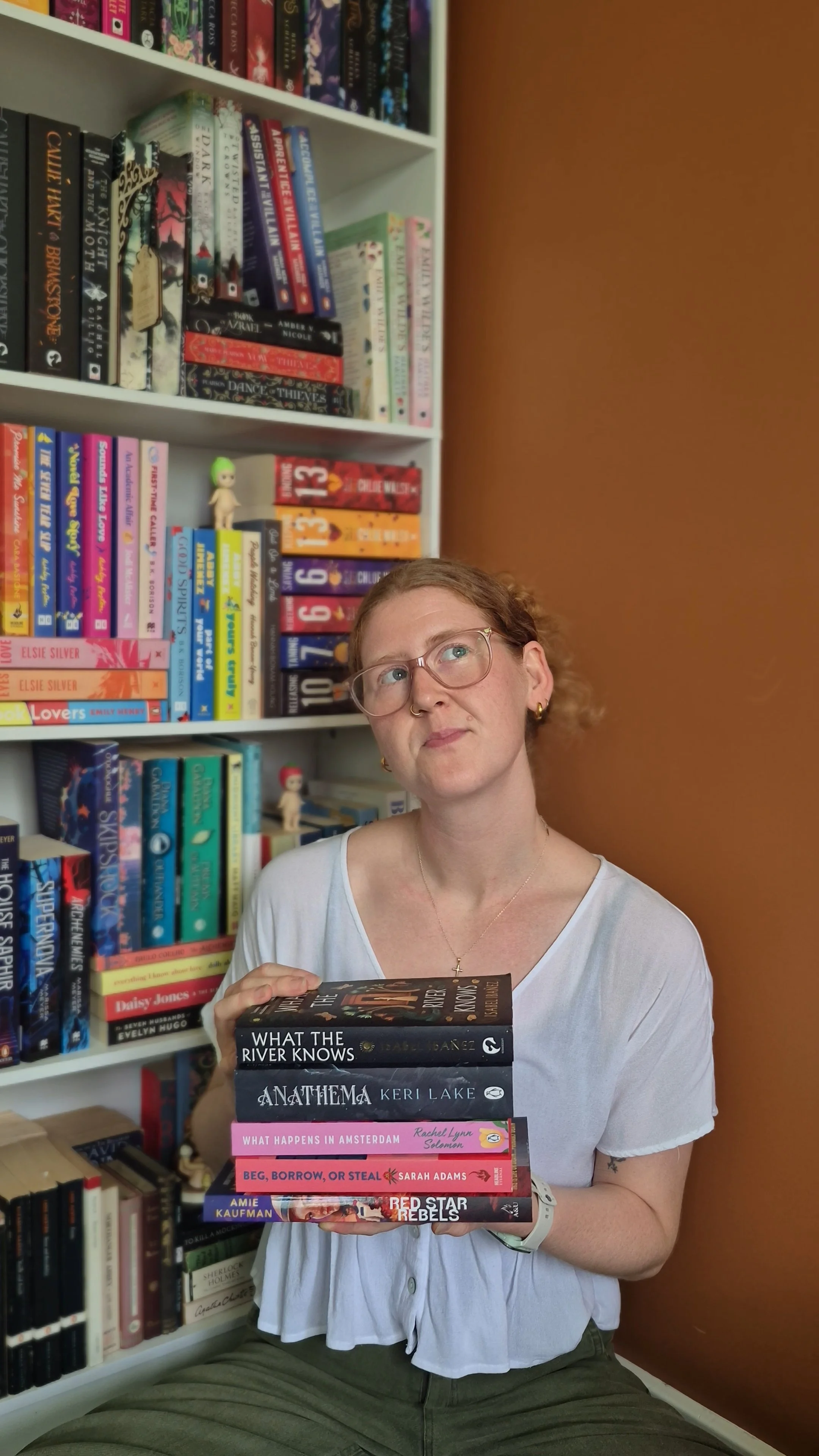 A woman with red hair in a claw clip, glasses, and a white shirt holding a stack of books in front of a bookshelf filled with colourful books. The woman is seated against a rust/orange toned wall.