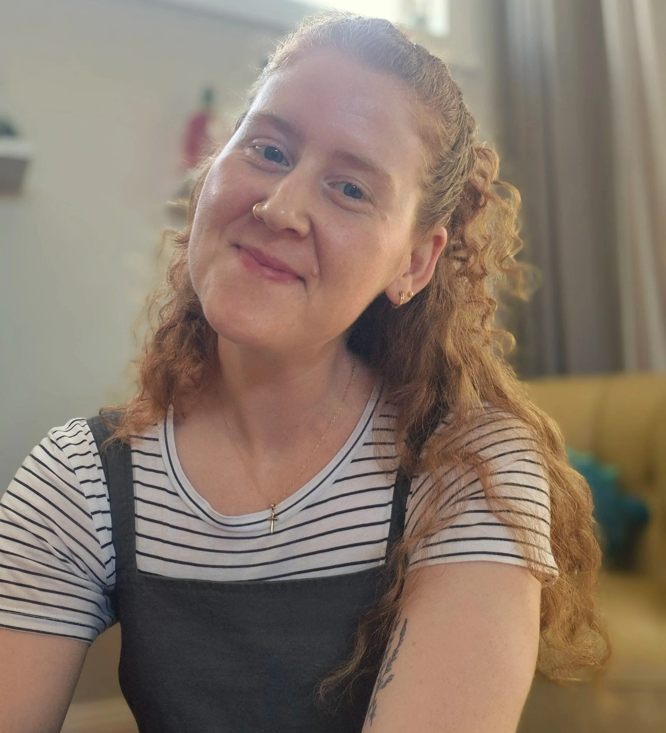 Close-up of a smiling woman with red curly hair, wearing a striped shirt in a well-lit indoor setting.