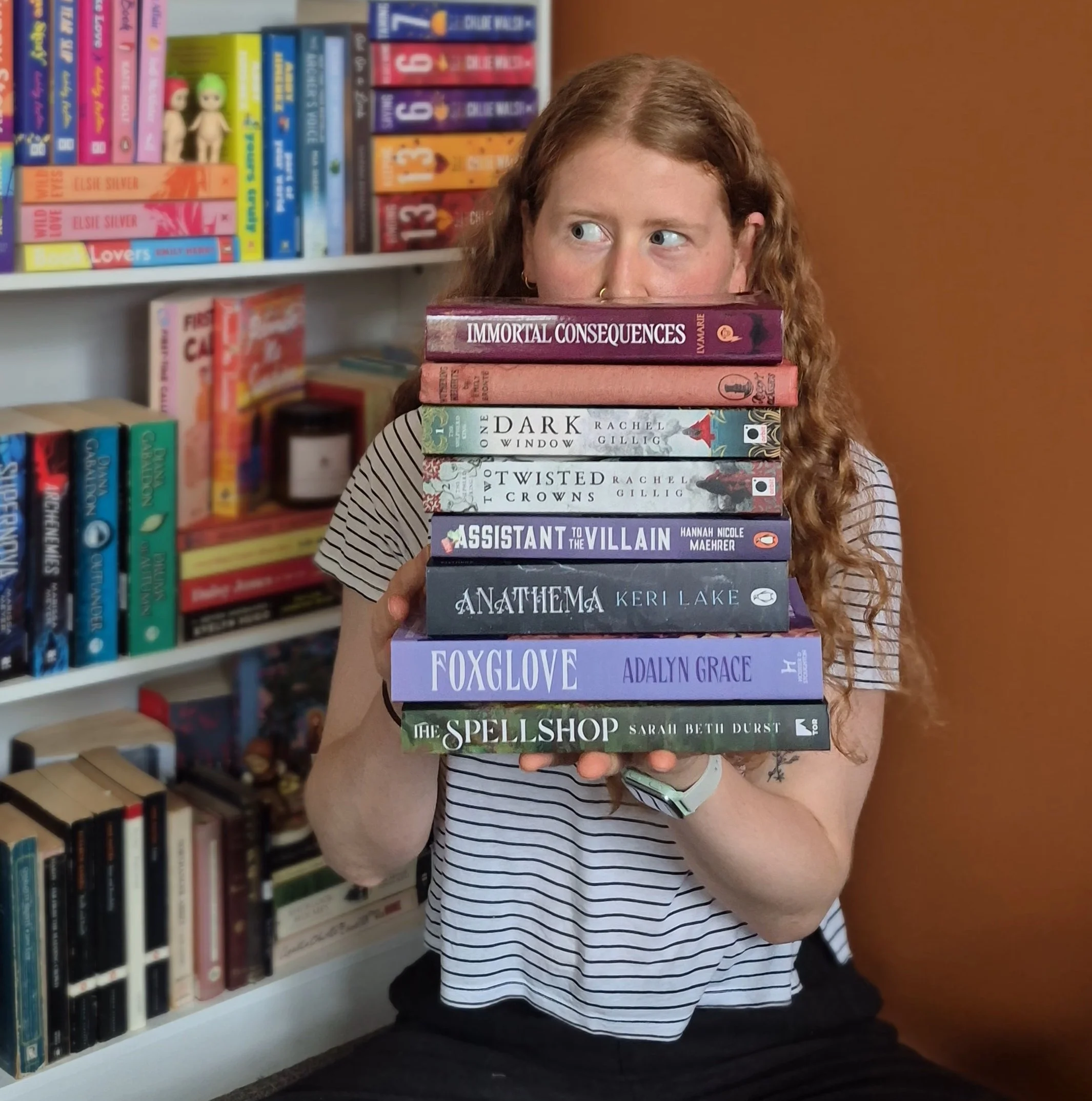 Person with long curly red hair holding a stack of books in front of their face in a library, with bookshelves filled with colorful books in the background.