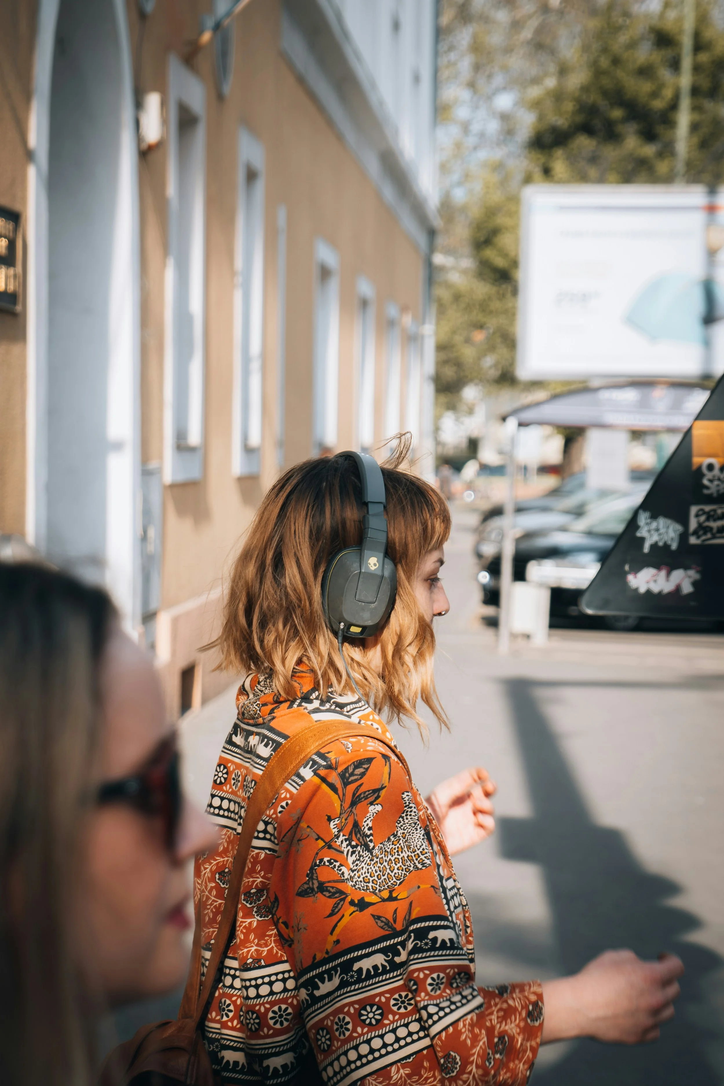 A person wearing headphones and a patterned orange and black jacket, standing outdoors on a sidewalk on a sunny day. They are facing to the right side.