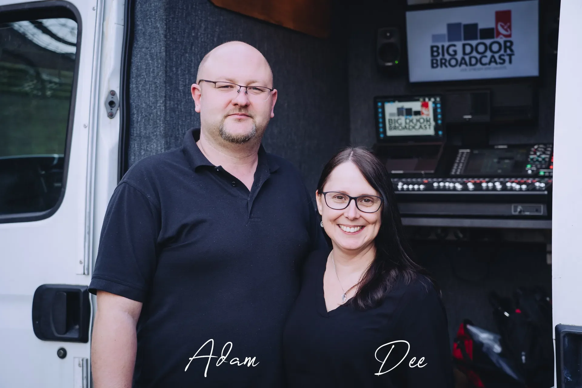 A man and woman standing outside of a vehicle with a broadcast setup inside, including a monitor and speaker, and a sign reading "Big Door Broadcast" in the background.