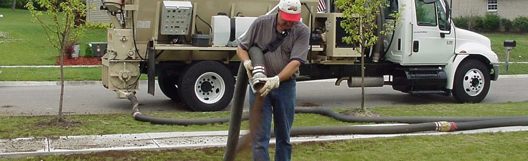 A worker is holding a large hose connected to a vacuum truck on a suburban street, with a lawn and small trees in the background.