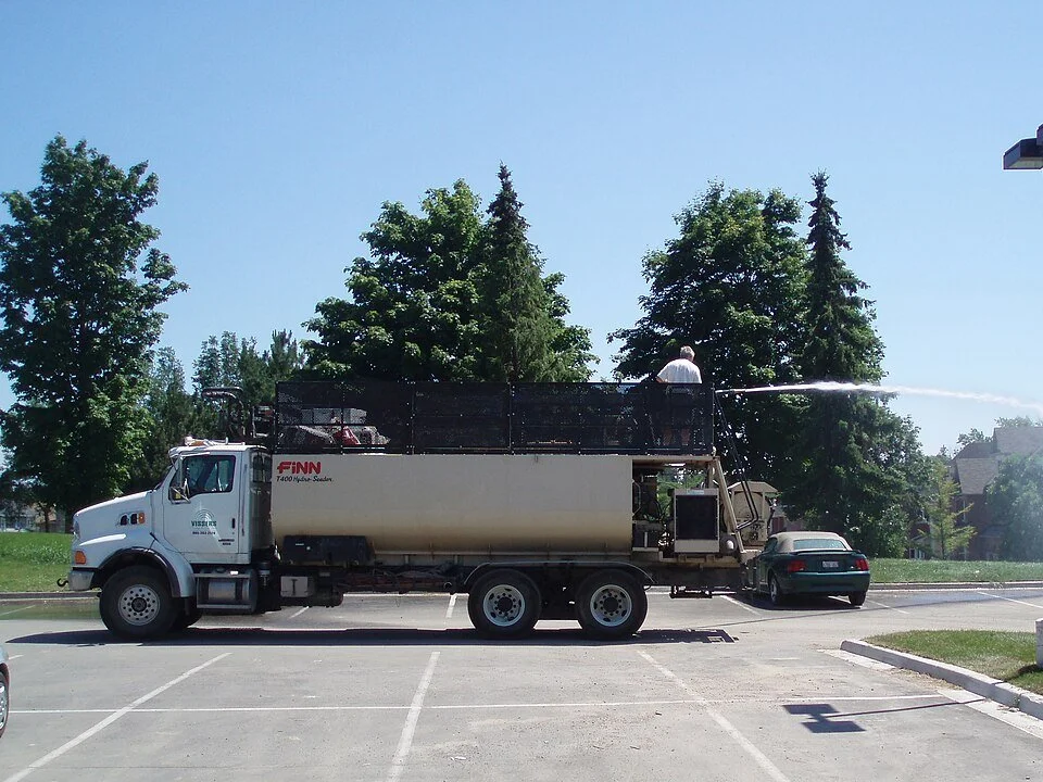 A water truck spraying water in a parking lot near trees with a person standing on the truck's platform.