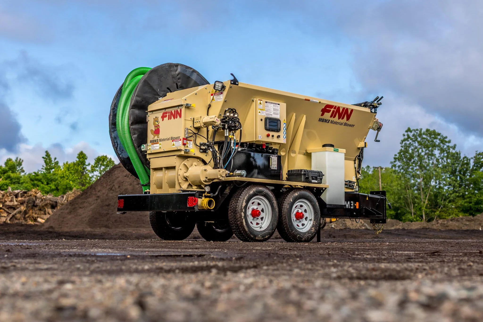 A large industrial machine labeled 'Finn MBM3 Material Blower' is stationed outdoors on a dirt surface, with a pile of soil or compost behind it, green trees in the background, and a partly cloudy sky overhead.