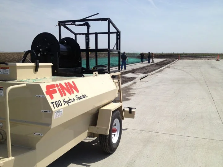 Agricultural seeder machine labeled 'FINN T60 Hydro Seeder' parked on a concrete surface with people in the background near a field under a clear sky.