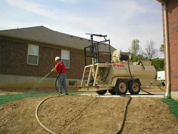 A worker in a red shirt and white helmet watering a lawn near a construction trailer in a backyard with a two-story brick house.