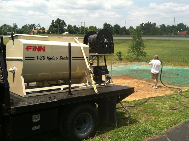 Man spraying water on a field with a hose near a large white water tank labeled 'FINN T-30 Hydro Seeder' on a trailer, with trees and a road in the background.