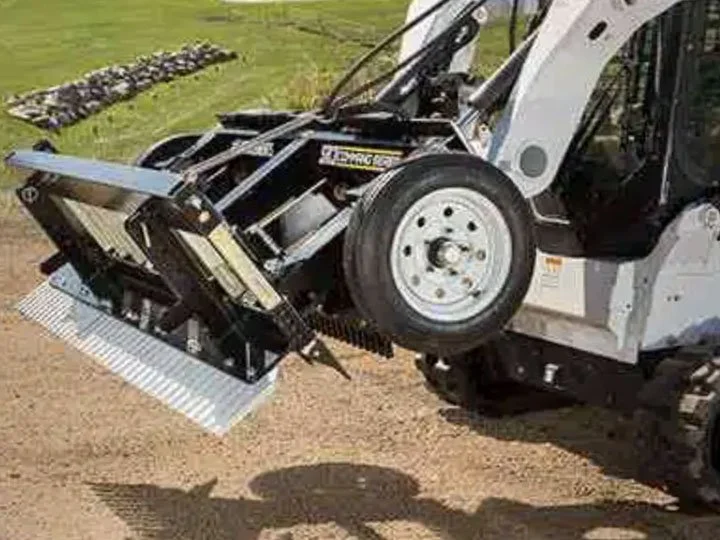 Close-up of a front loader attachment on a construction or landscaping vehicle, with a gravel road in the background.