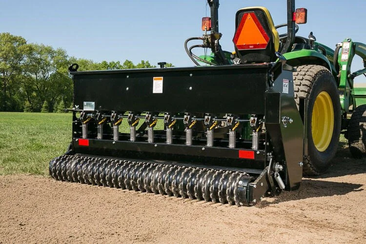 A green tractor with a large black and yellow attachment used for aerating soil on a farm field.
